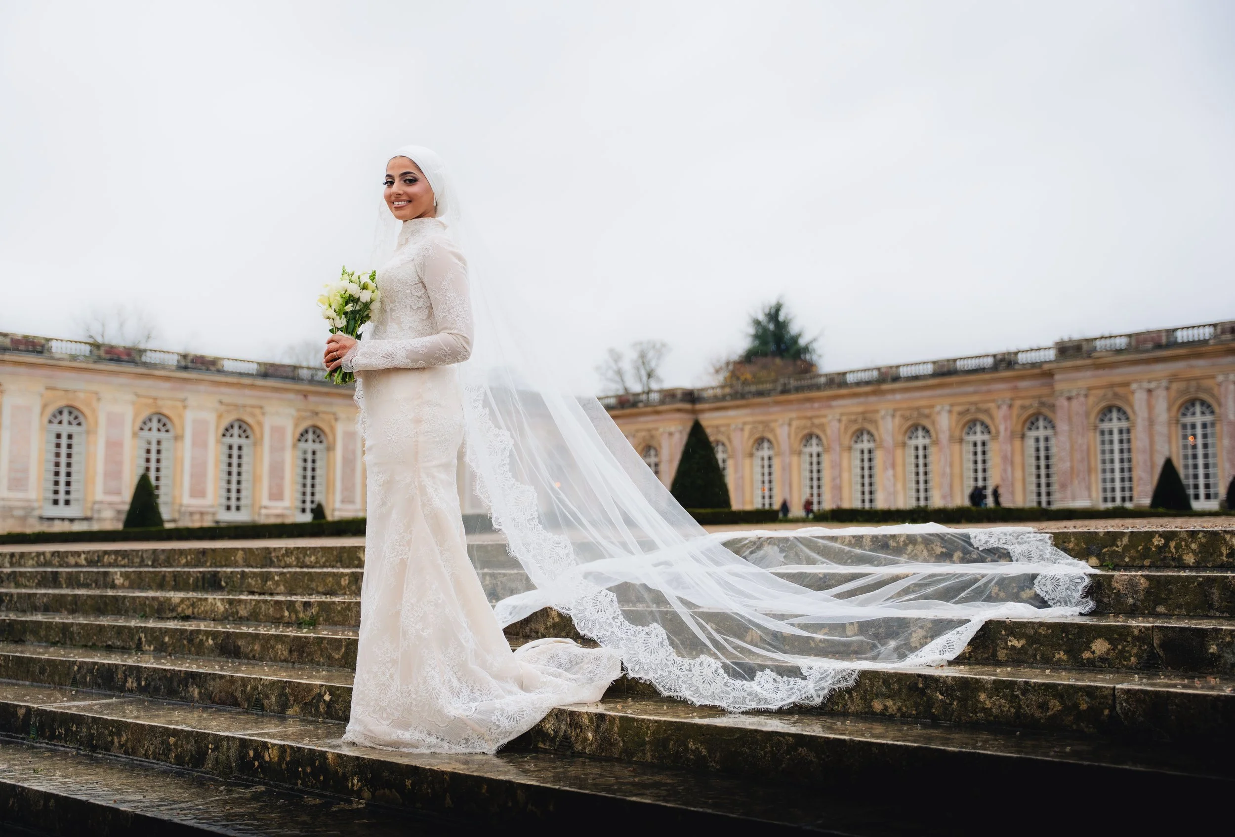 A smiling bride in a white lace wedding gown holding a bouquet of white flowers, standing on stone steps outdoors with a historic building in the background and overcast sky.