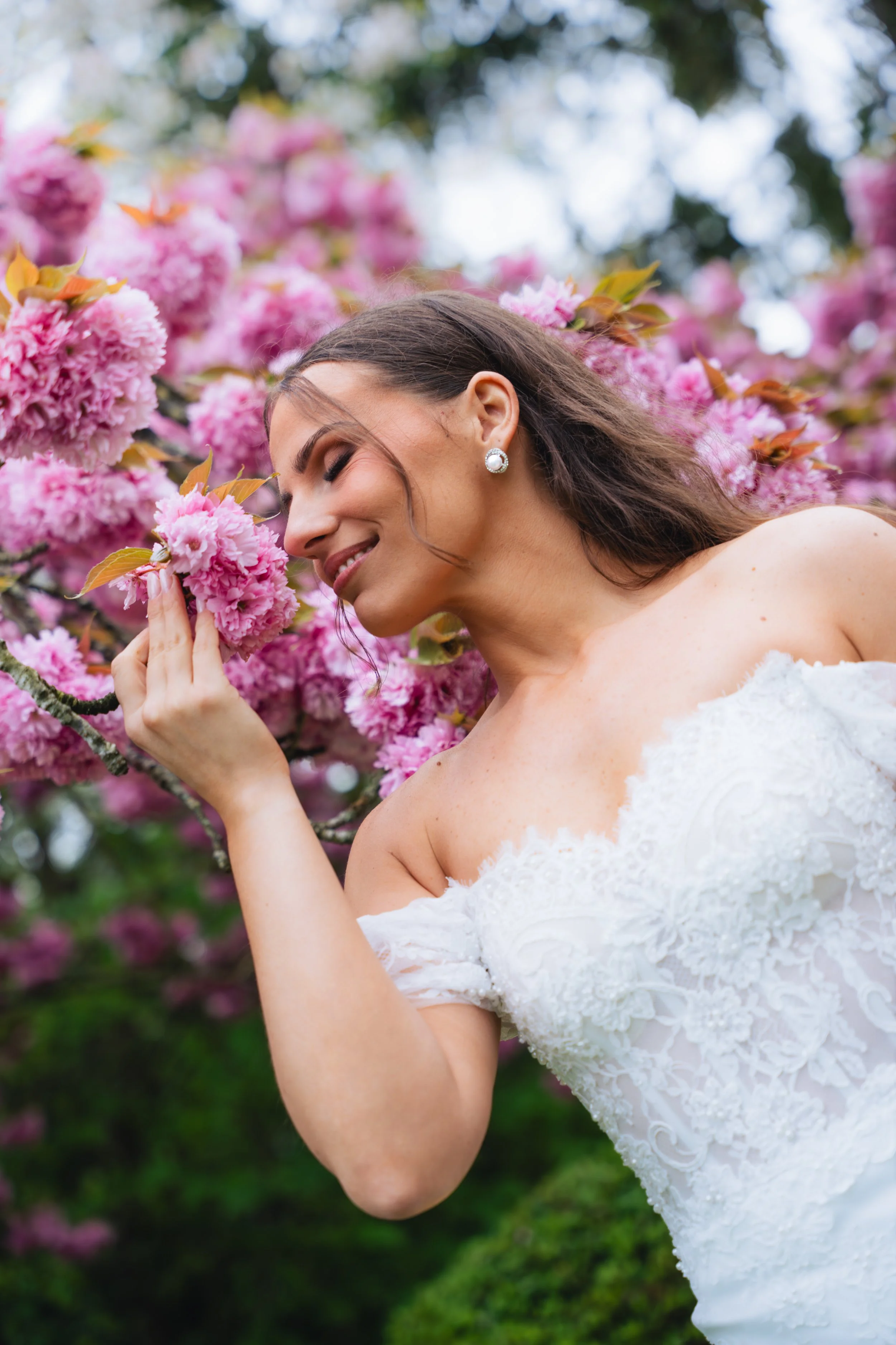A woman in a white wedding dress smiling with her eyes closed, holding a pink flower close to her nose, surrounded by pink blossoms and green leaves.