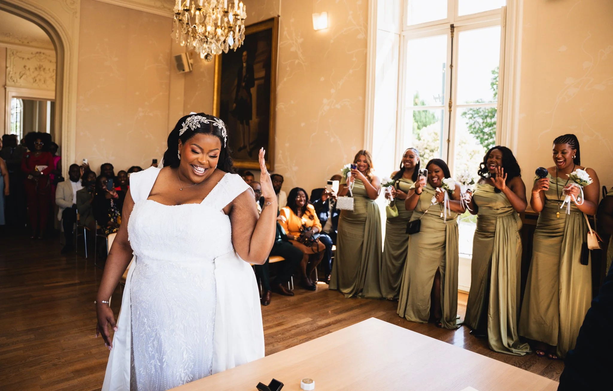 A bride in a white wedding dress throws her bouquet into the air as a group of bridesmaids and guests cheer and smile in a brightly lit room with large windows and a chandelier.