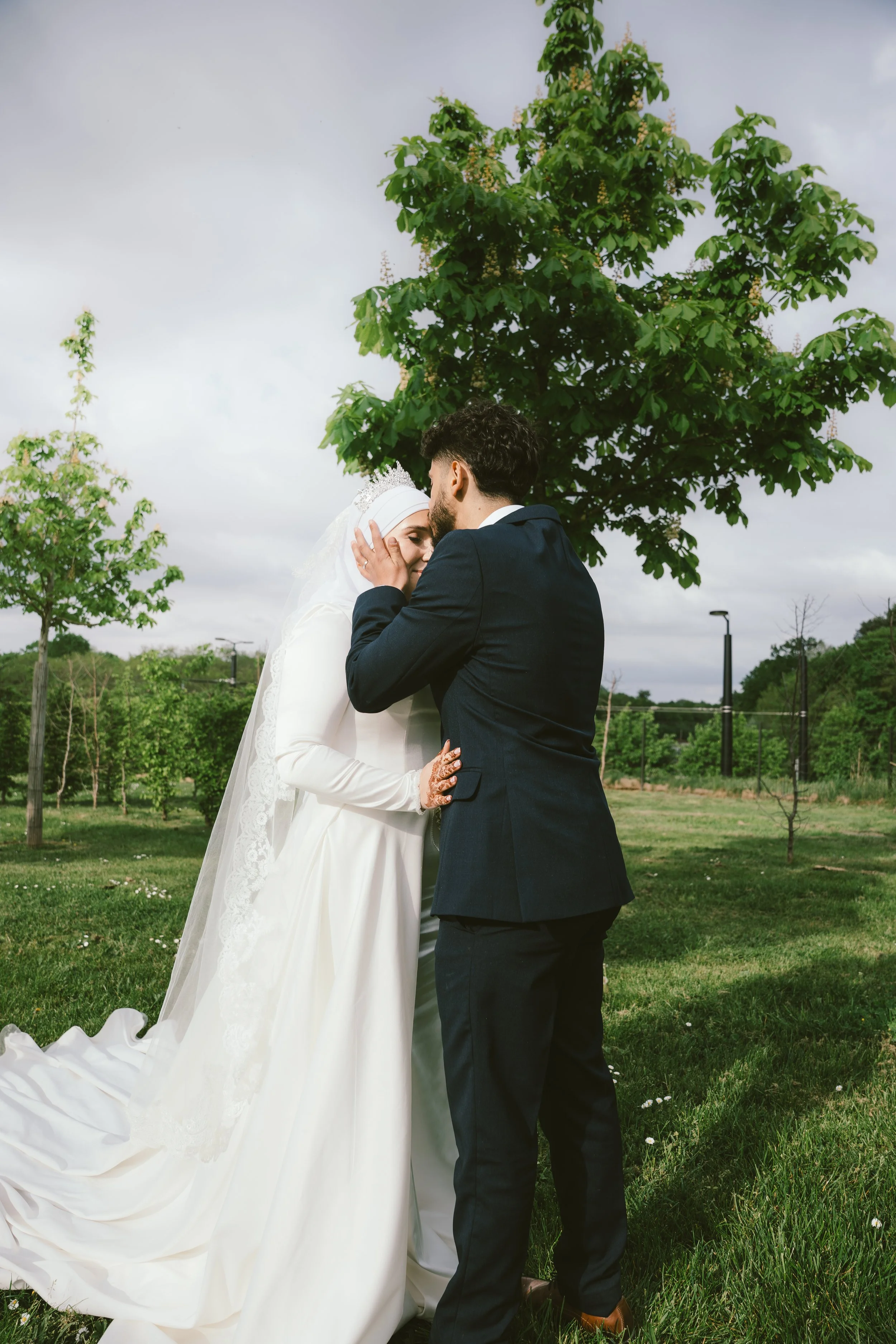 Un couple de mariés échange un baiser dans un parc verdoyant sous un ciel nuageux. La mariée porte une robe blanche longue avec un voile et une couronne, le marié porte un costume sombre. Ils se tiennent près d'un grand arbre vert.