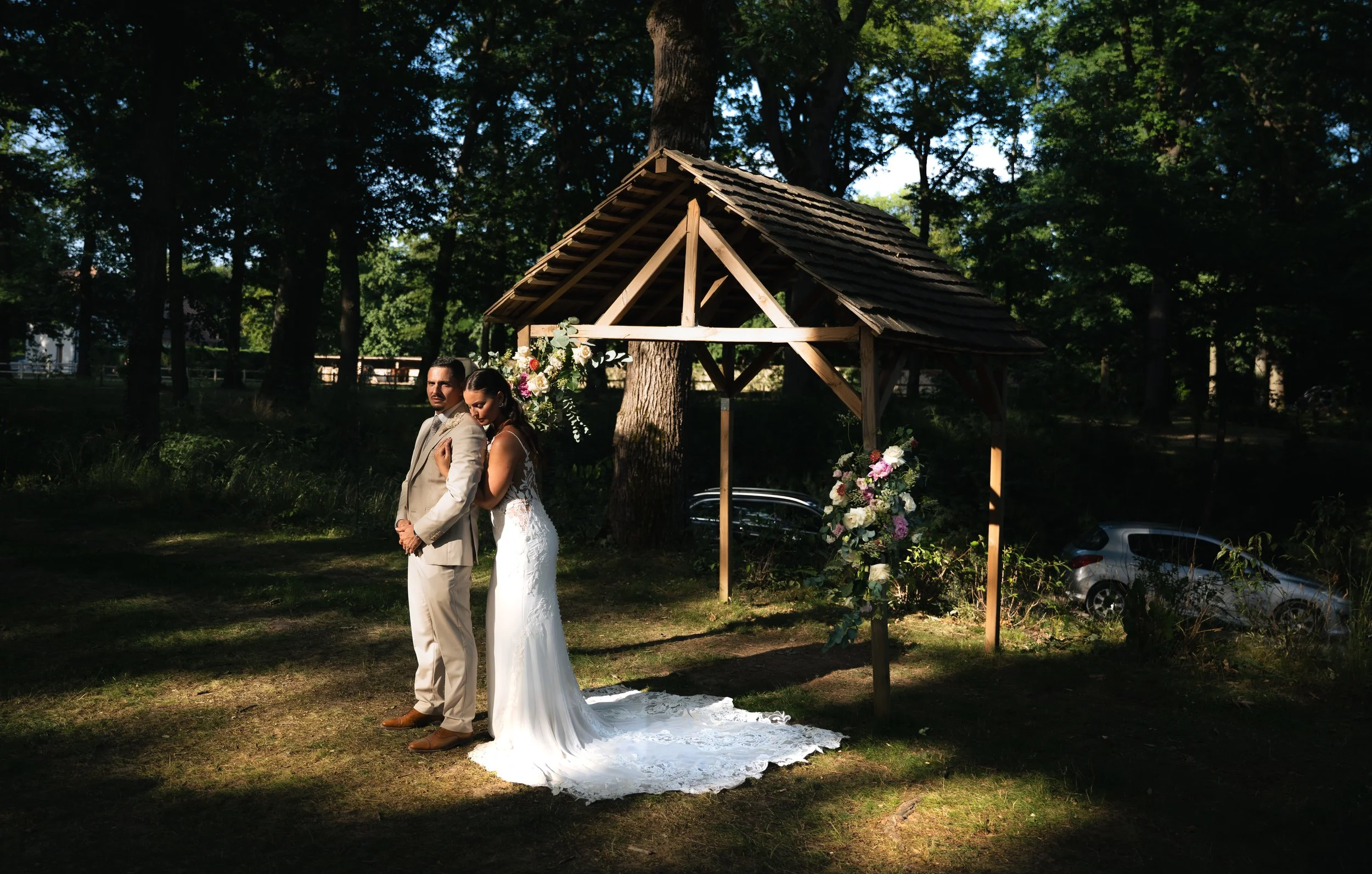 A bride and groom stand together outdoors during a wedding ceremony in a wooded area. The bride is wearing a white lace dress and the groom is wearing a light-colored suit. They are standing under a wooden wedding arch decorated with flowers.