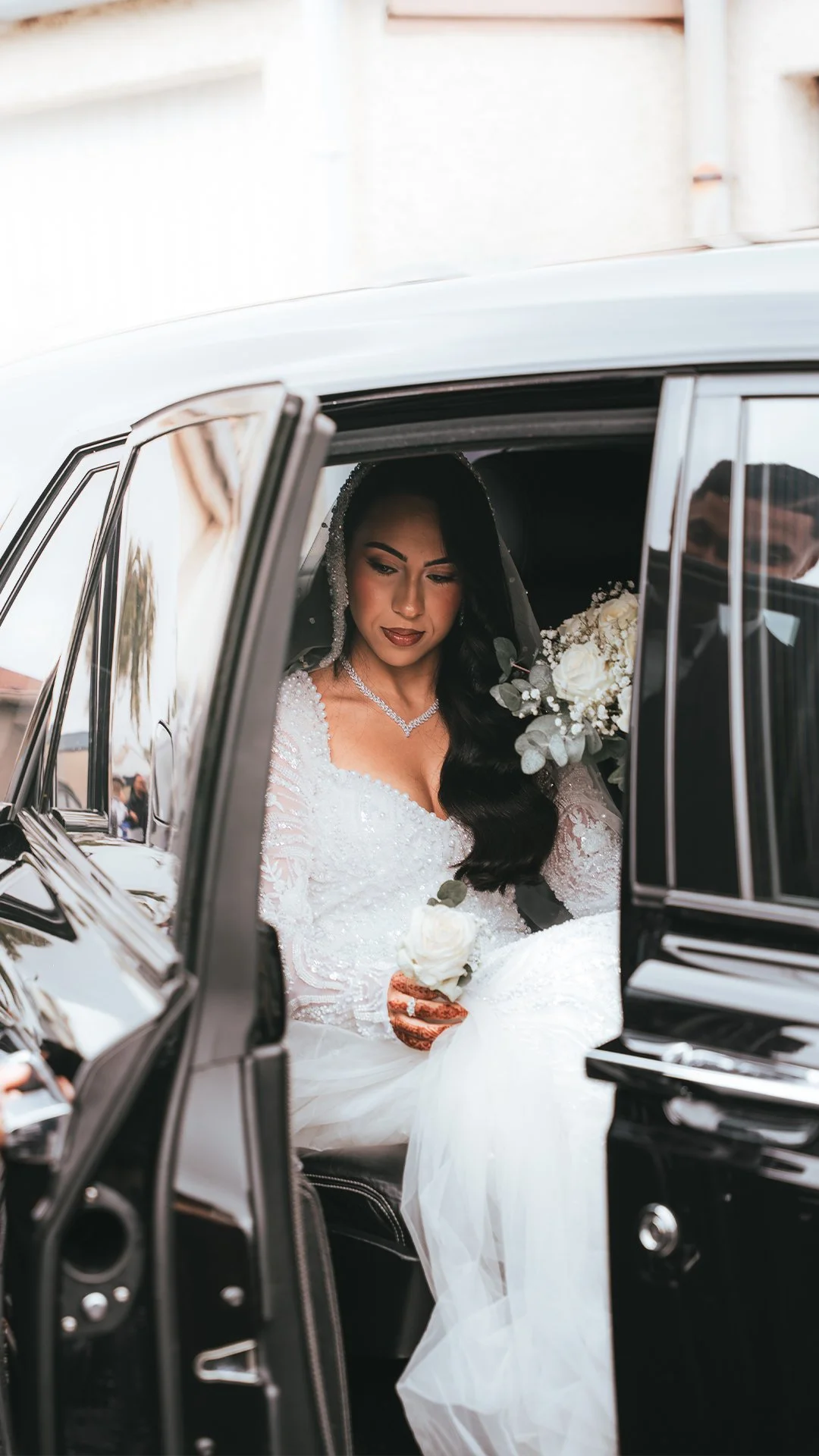 Bride in white wedding dress sitting in a vintage black car holding a bouquet of white roses and greenery, with a man partly visible behind her.