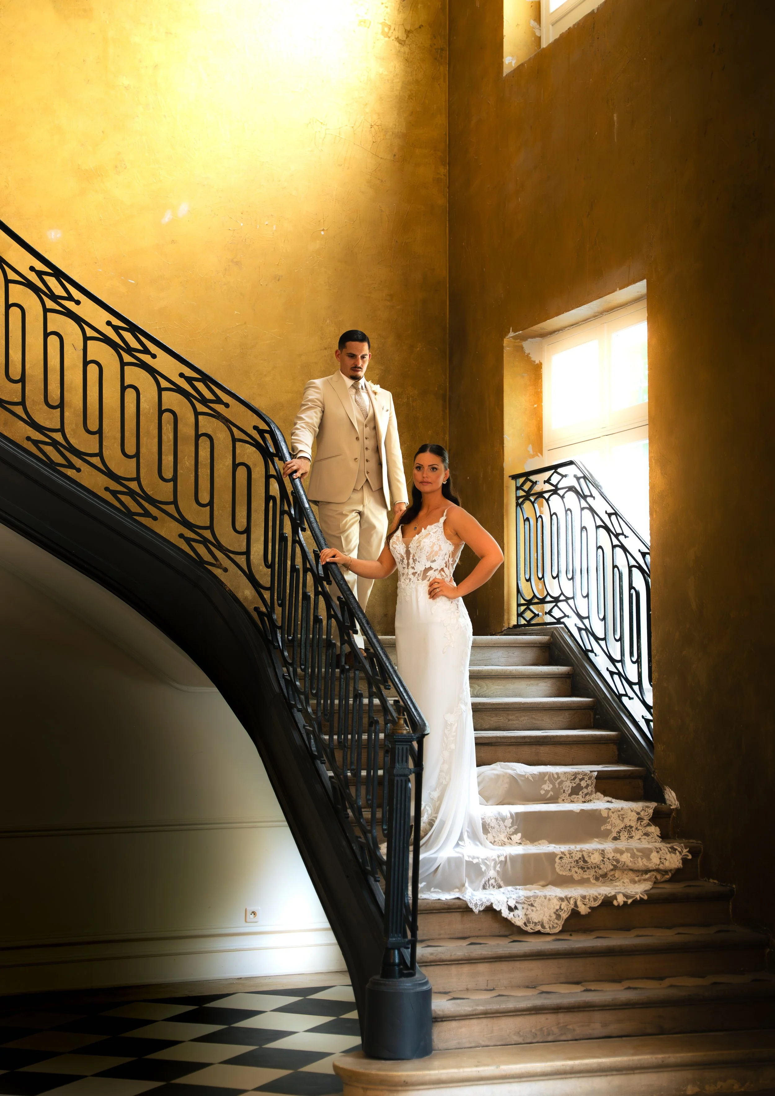 A bride and groom standing on a staircase in an elegant, historic building with tall windows and warm yellow walls. The bride wears a white lace wedding gown with a train, and the groom is dressed in a cream-colored suit. They pose in a formal, roman