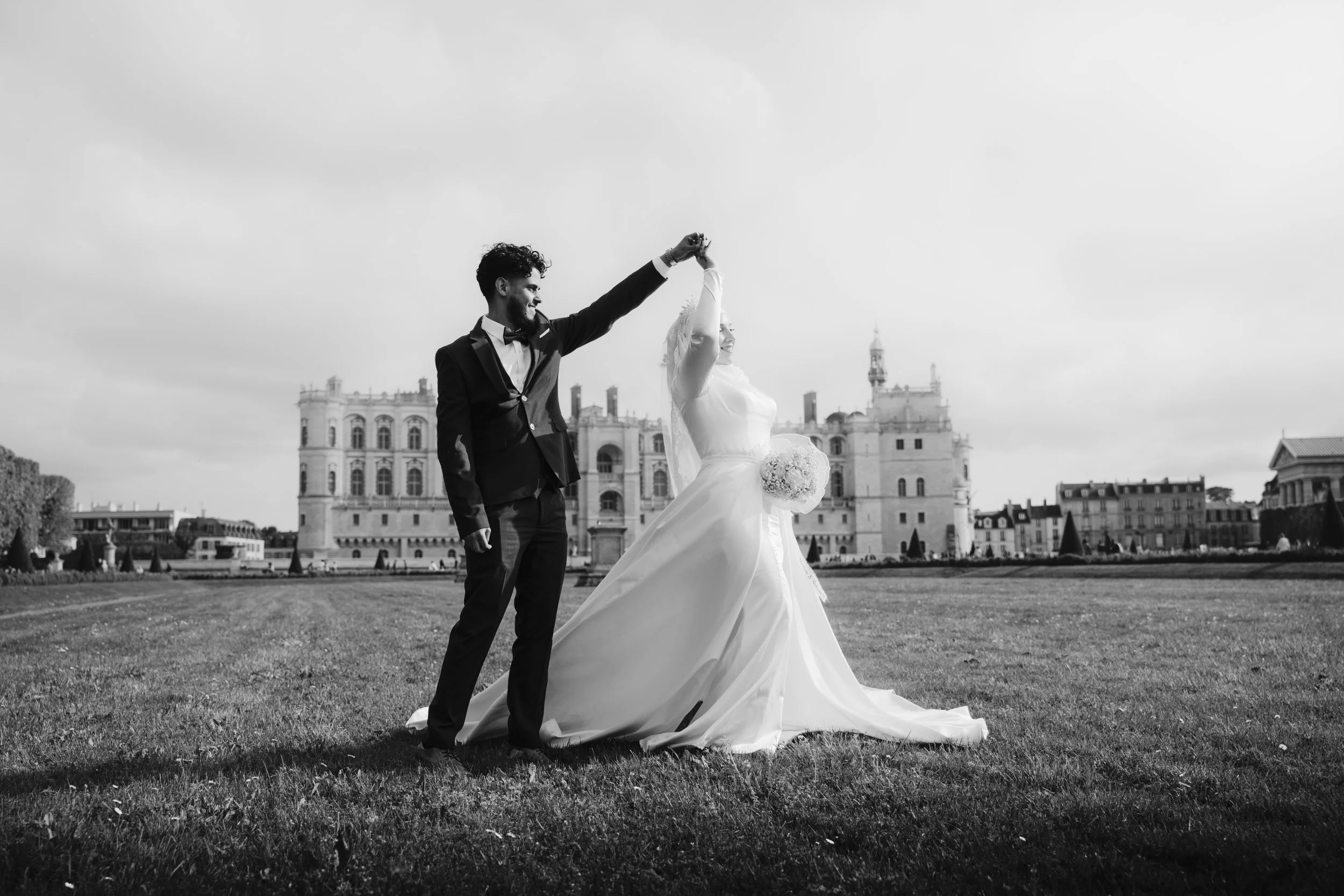 Un couple marié danse dans un parc devant un grand bâtiment historique, la photo est en noir et blanc.