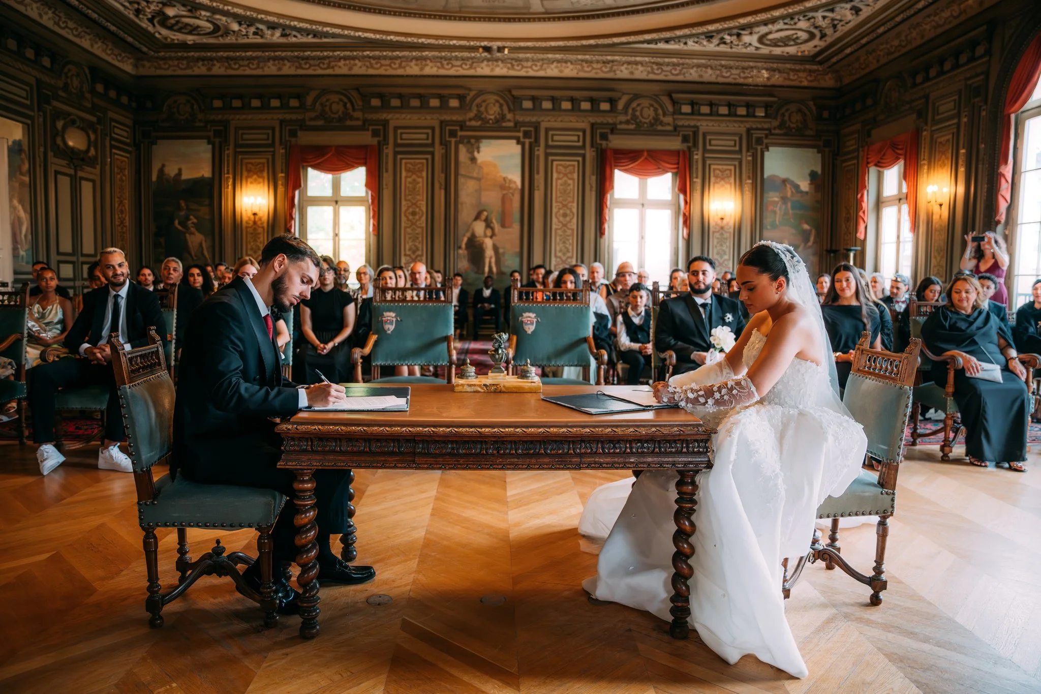 A wedding ceremony taking place in a grand, ornate room with high ceilings, large windows, and painted walls. The bride and groom are seated at a table, with the bride in a white wedding dress and the groom in a dark suit. Guests are seated behind th
