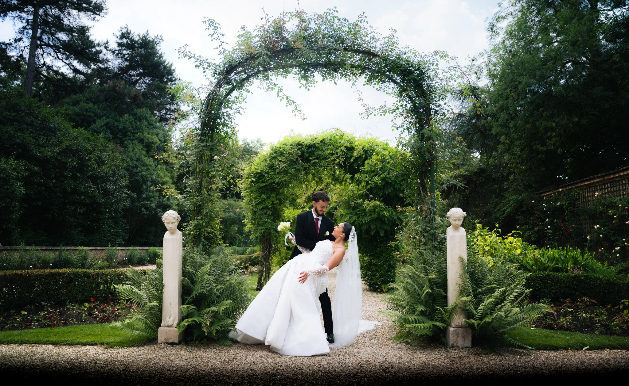 A bride and groom on their wedding day in a lush green garden with statues and an arched trellis overhead.