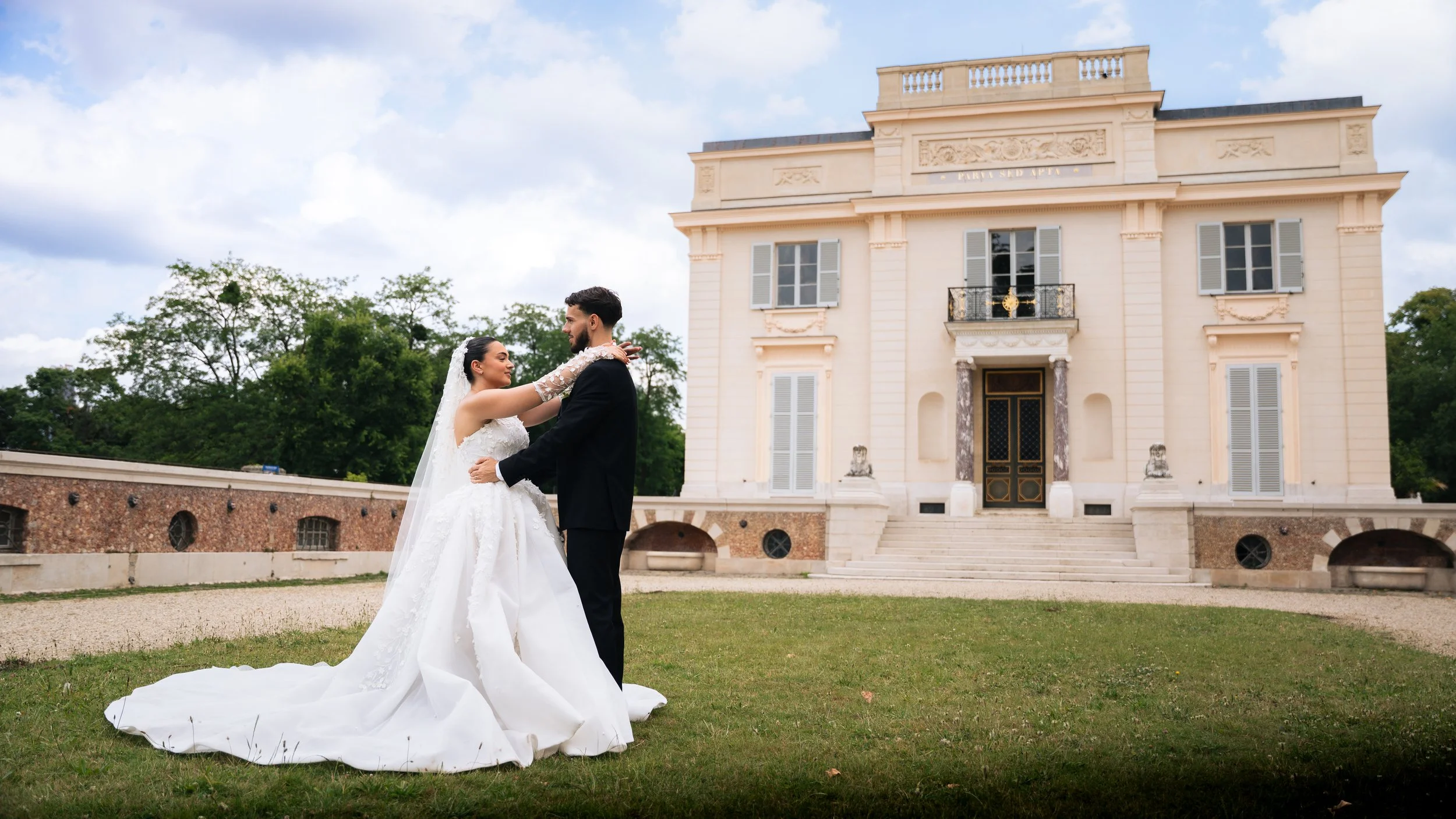 A bride and groom dance outside in front of a large, elegant, cream-colored mansion with multiple windows and a staircase leading to the main entrance.