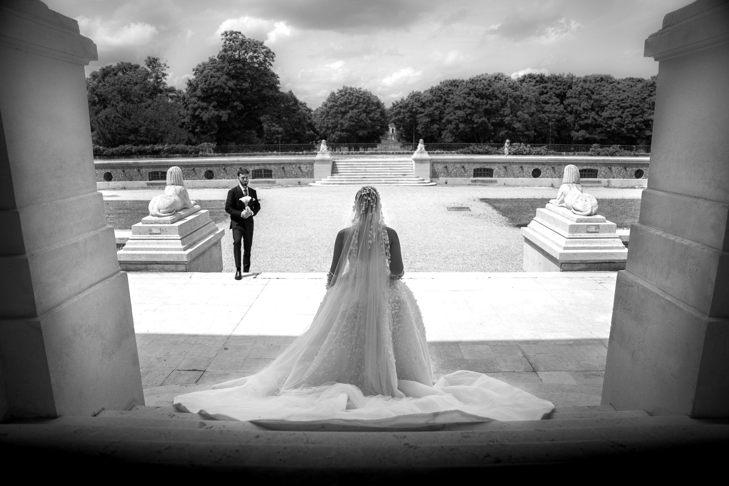 A bride in a wedding dress with a train standing on steps viewing a groom in a tuxedo holding a bouquet while facing each other outdoors at a formal wedding ceremony, framed by stone columns and lion statues, with trees and a cloudy sky in the backgr
