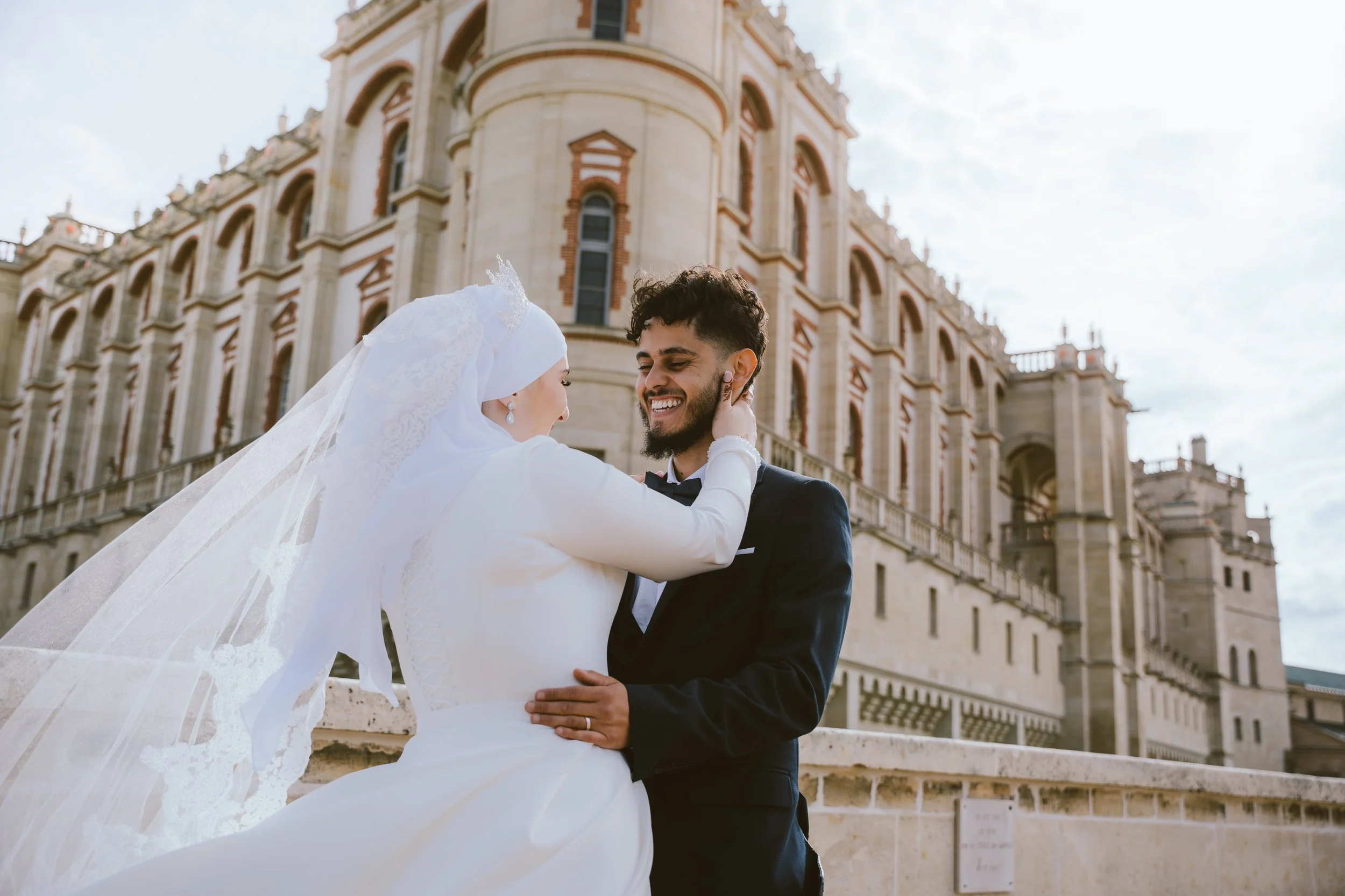 Un couple en mariage en tenue élégante, souriant et se regardant tendrement devant un bâtiment historique en pierre, avec un ciel partiellement nuageux.