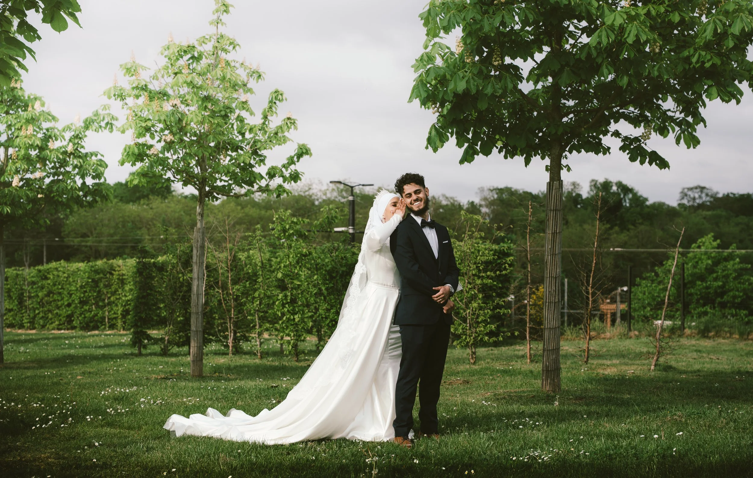 Un couple de mariés posant dans un parc, entouré d'arbres verts. La femme porte une robe de mariée blanche avec une longue traîne, et l'homme porte un costume noir avec une cravate noire, tous deux souriants.