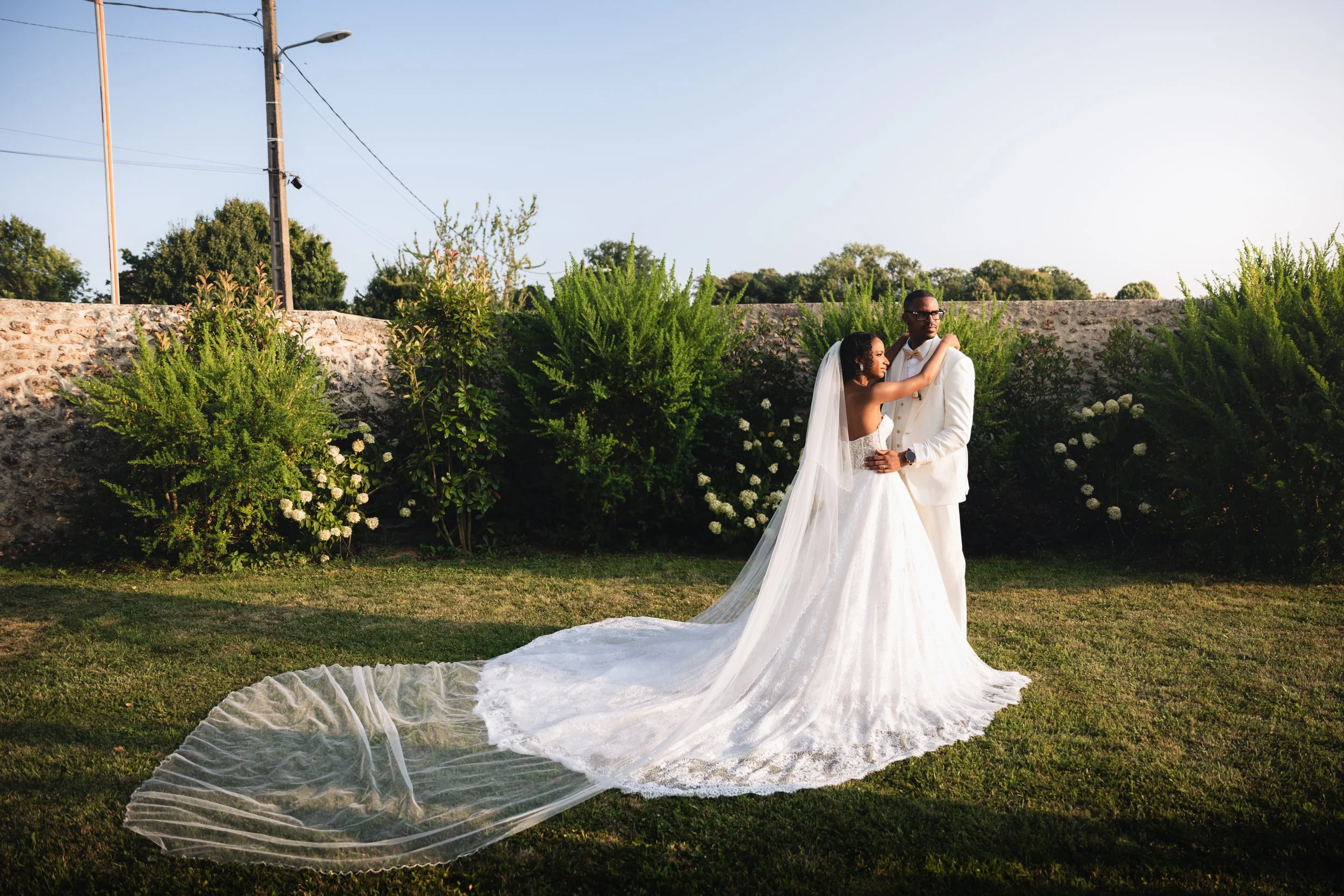 A bride and groom standing together outdoors, the bride wearing a white wedding gown and veil and the groom wearing a white tuxedo, surrounded by greenery and plants, during a wedding photo shoot.