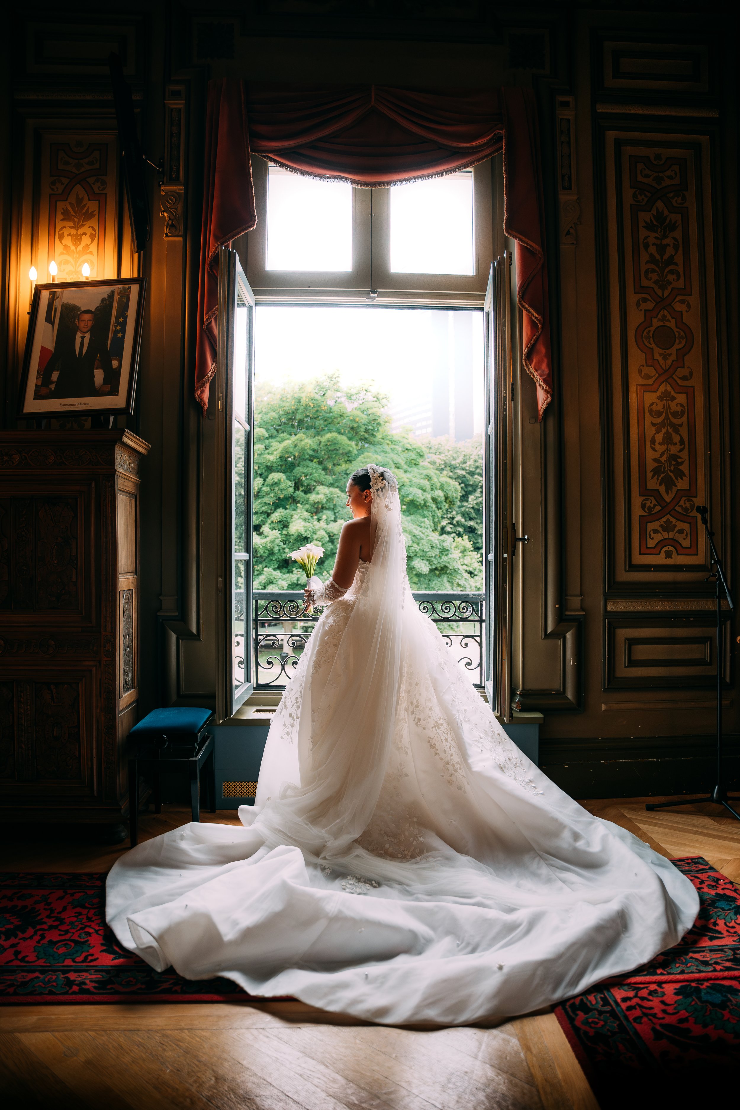 A bride in a white wedding gown holding a bouquet, standing near an open window in a decorated room with wooden panels and a framed portrait on the wall.