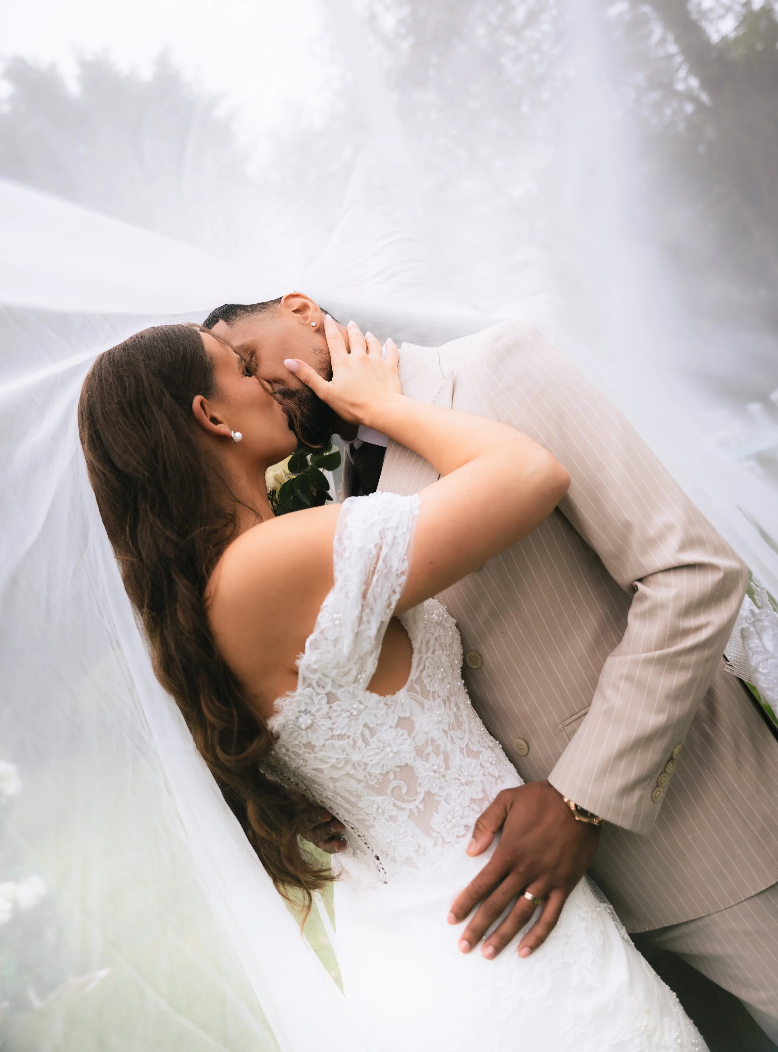 A bride and groom sharing a kiss beneath a white veil outdoors, with the bride in a lace wedding dress and pearl earrings, and the groom in a light-colored pinstripe suit.