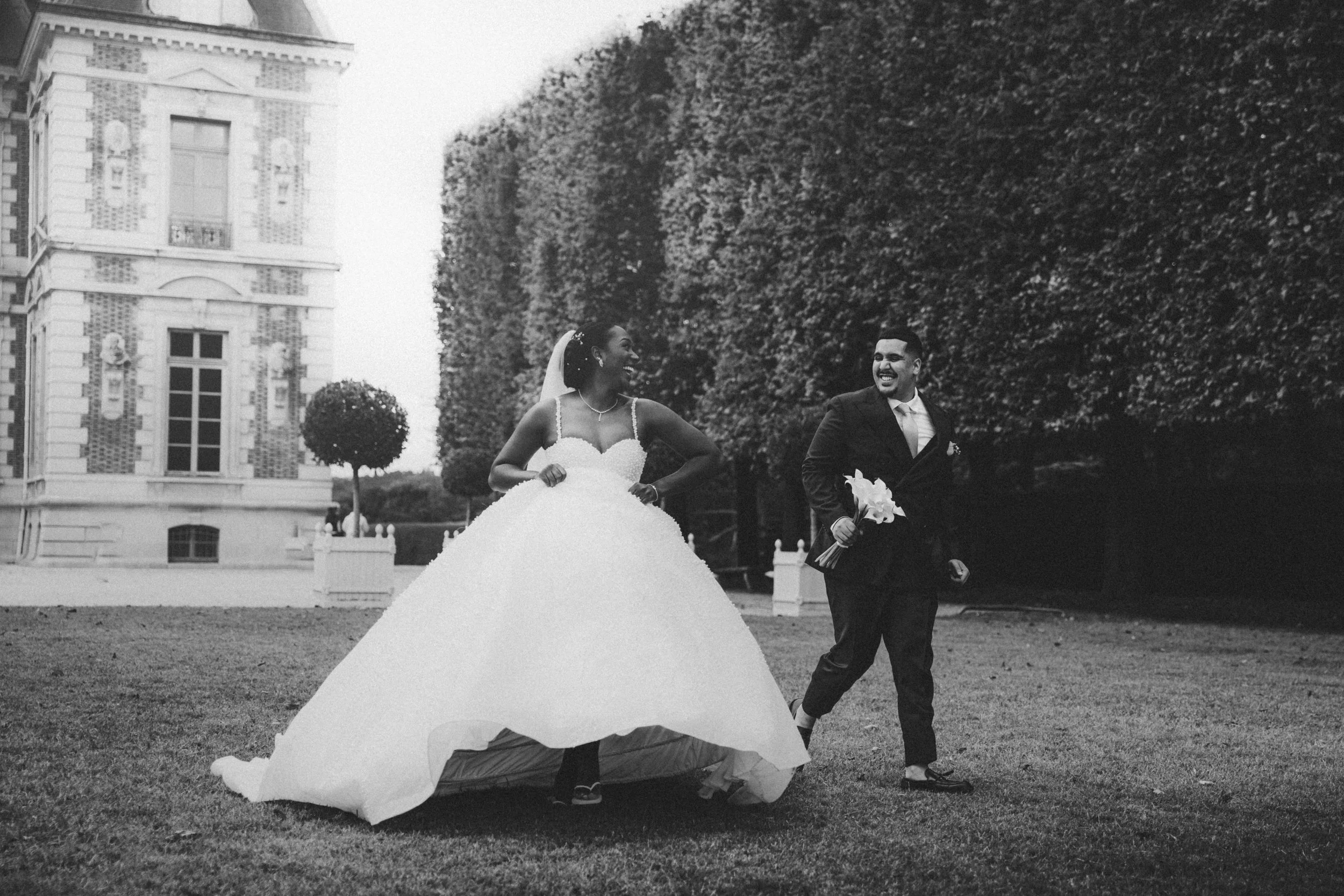 A black and white photo of a bride and groom outdoors in front of a large building with trees and bushes. The bride is smiling and wearing a wedding dress, while the groom, holding a bouquet, is smiling and walking beside her.