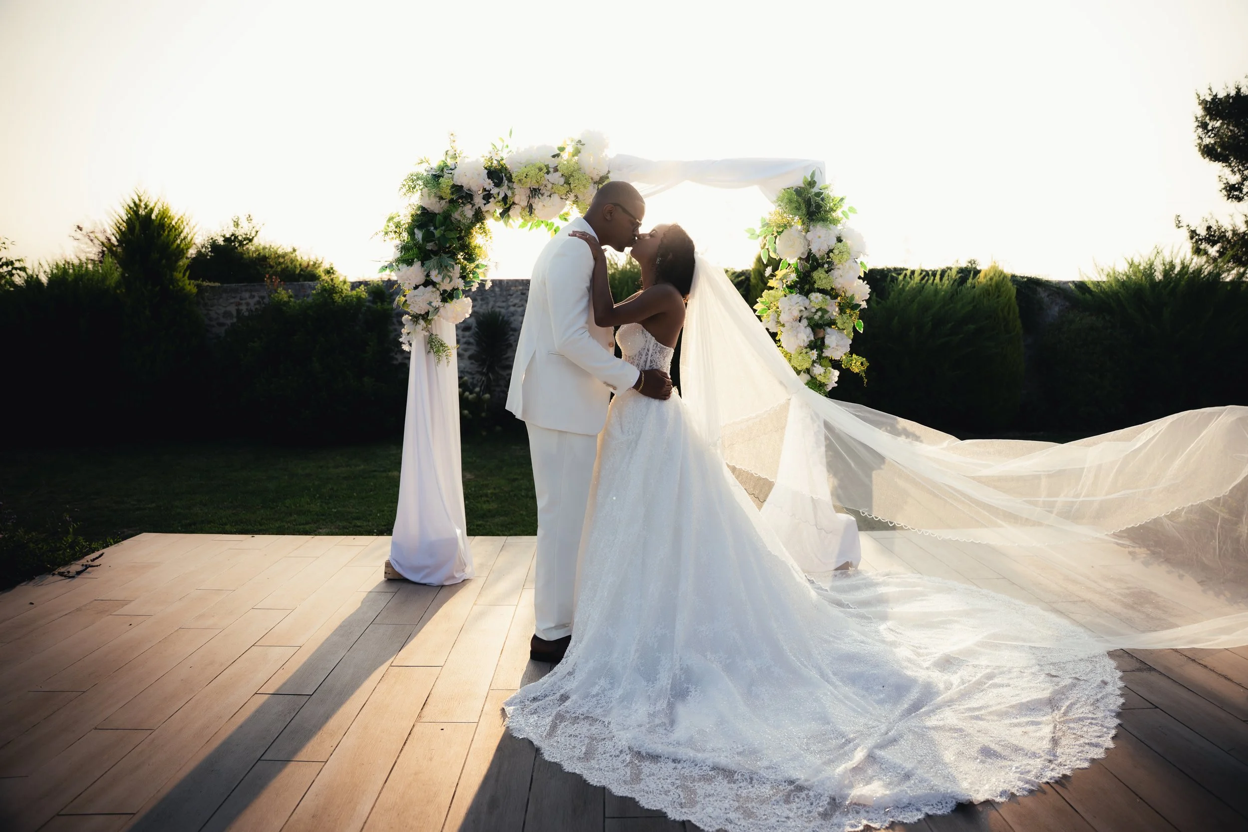 A newlywed couple shares a kiss under a floral arch at sunset, outdoors, with trees and a stone wall in the background.
