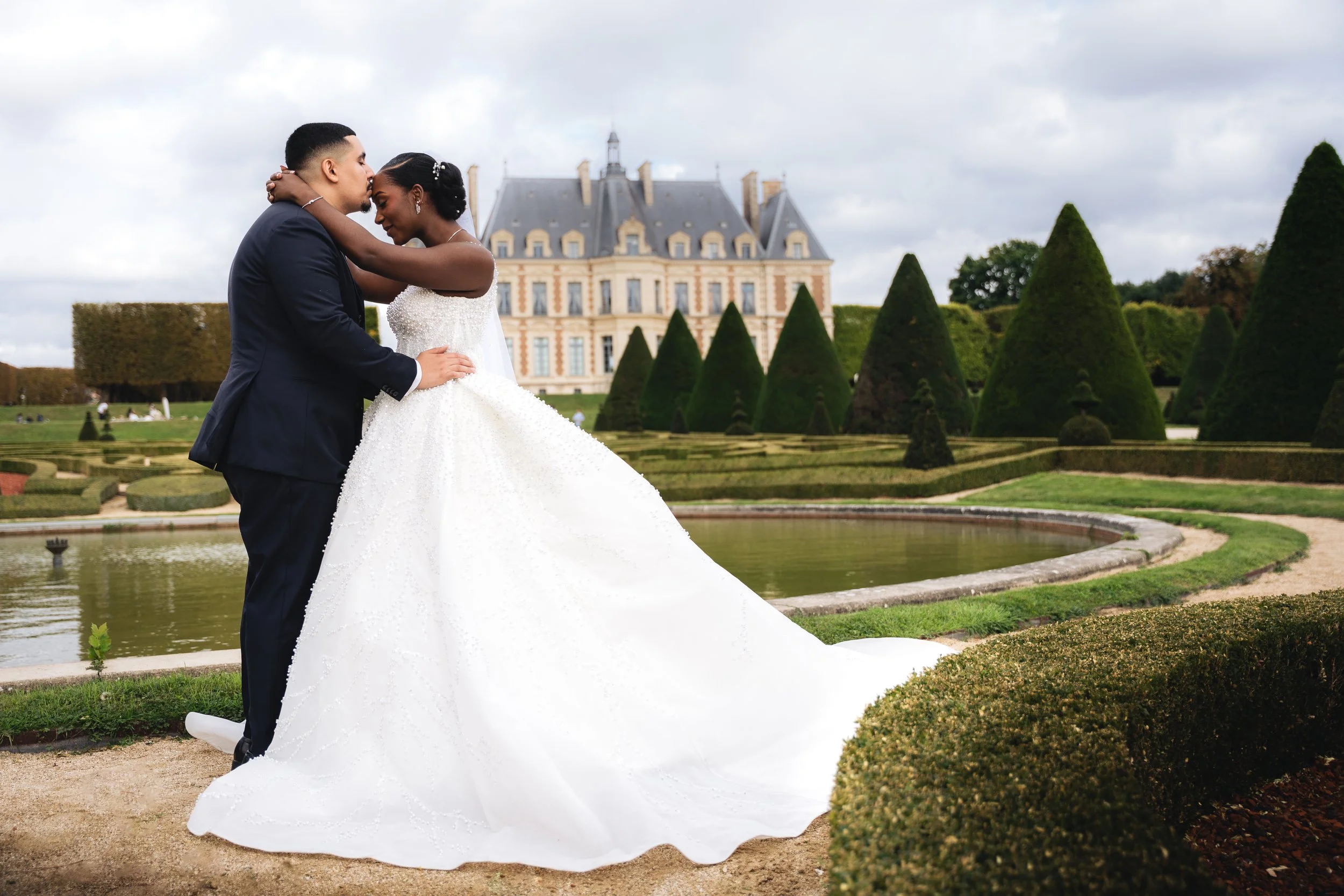 A bride and groom embracing in front of a French chateau-style building with landscaped gardens.