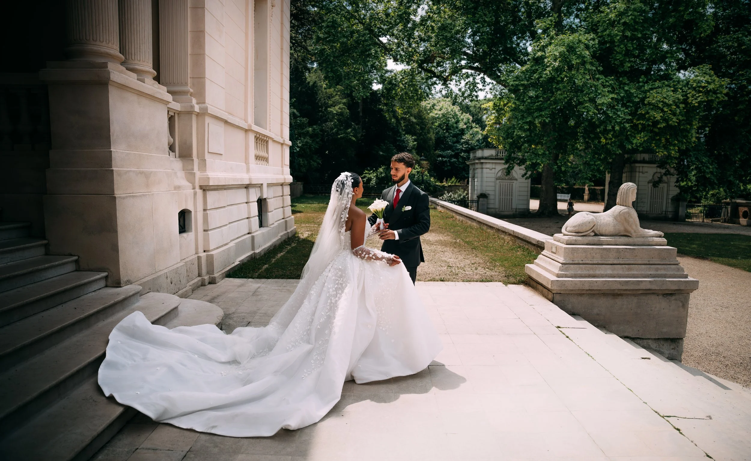 A bride and groom standing outdoors in front of a historic building, with large trees in the background. The bride wears a white wedding gown and veil, holding a bouquet, and the groom dressed in a dark suit with a red tie. They are looking at each other during their wedding ceremony.