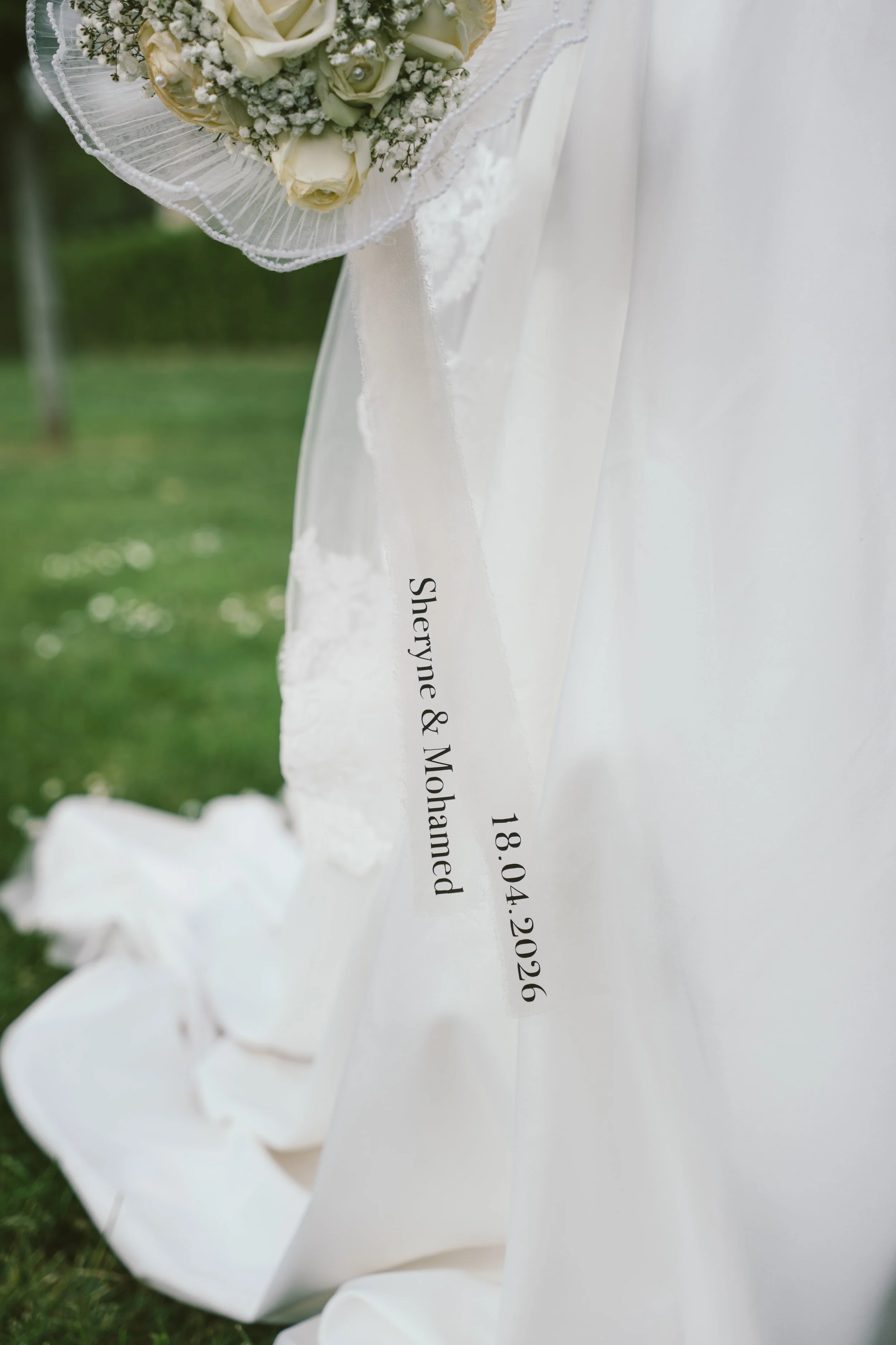 Un bouquet de fleurs blanches avec des roses et des œillets, enroulé dans une dentelle, posé à côté d'une robe de mariage blanche, portant une inscription partielle avec la date 18.04.2026.
