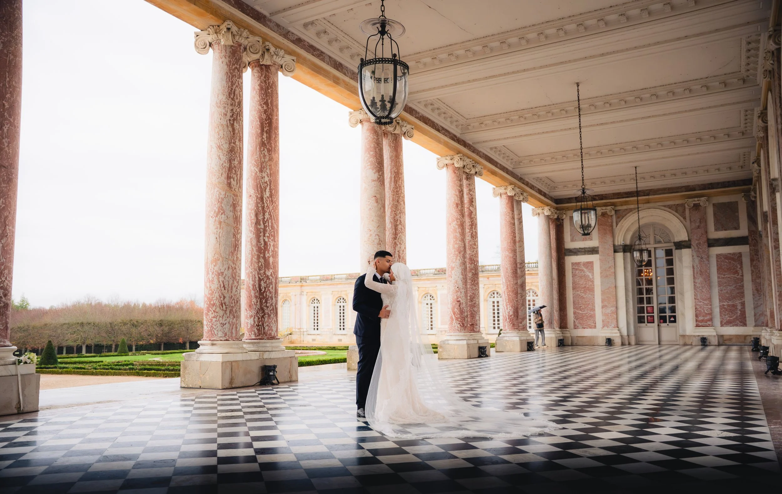 A bride and groom sharing a dance under a classical'', columned pavilion with ornate ceiling and hanging lanterns. The bride wears a white wedding gown, and the groom a tuxedo. The scene is set outdoors with a garden in the background.