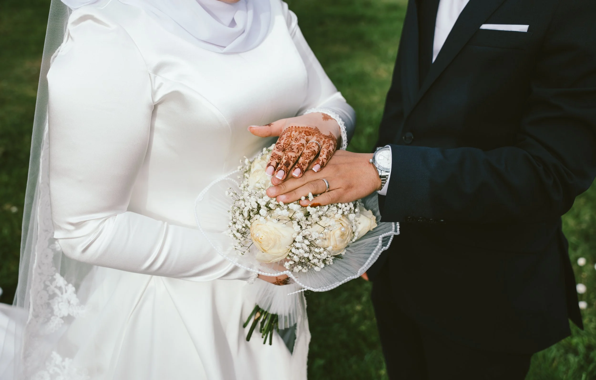 Close-up of a bride and groom during their wedding ceremony. The bride is holding a bouquet of white flowers, and her hand showcases intricate henna designs. She is wearing a white satin dress, and the groom is dressed in a black suit with a wristwatch.