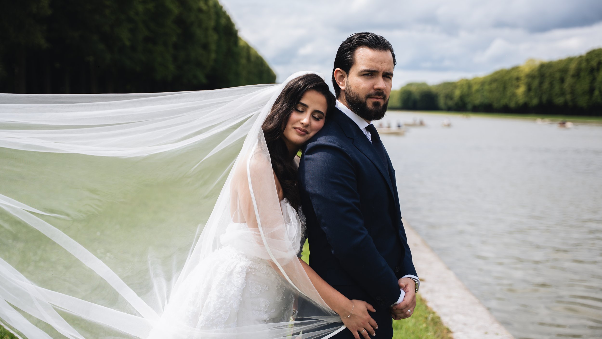 A bride and groom sitting by a river, with the bride leaning her head on the groom's shoulder and the groom looking at the camera. The bride wears a white wedding dress and a long veil, and the groom wears a dark suit. The background shows a cloudy sky, a river, and trees.