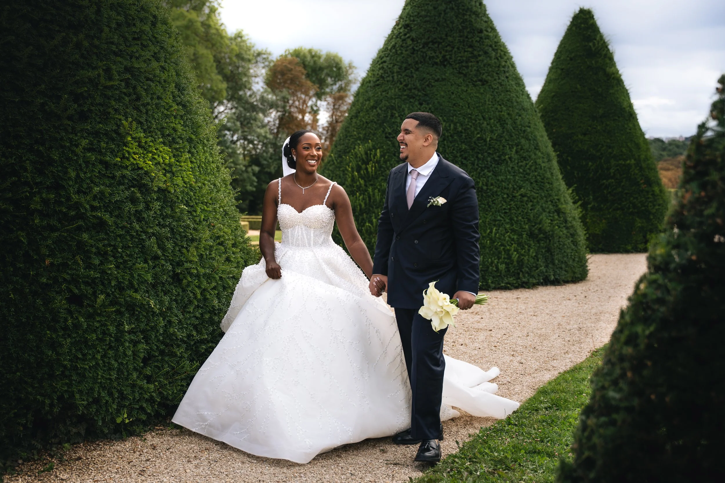 A newlywed couple holding hands and smiling while walking through a lush, green garden with tall, trimmed bushes and trees in the background. The bride is wearing a white wedding gown with beaded details and a veil, while the groom is in a dark suit 