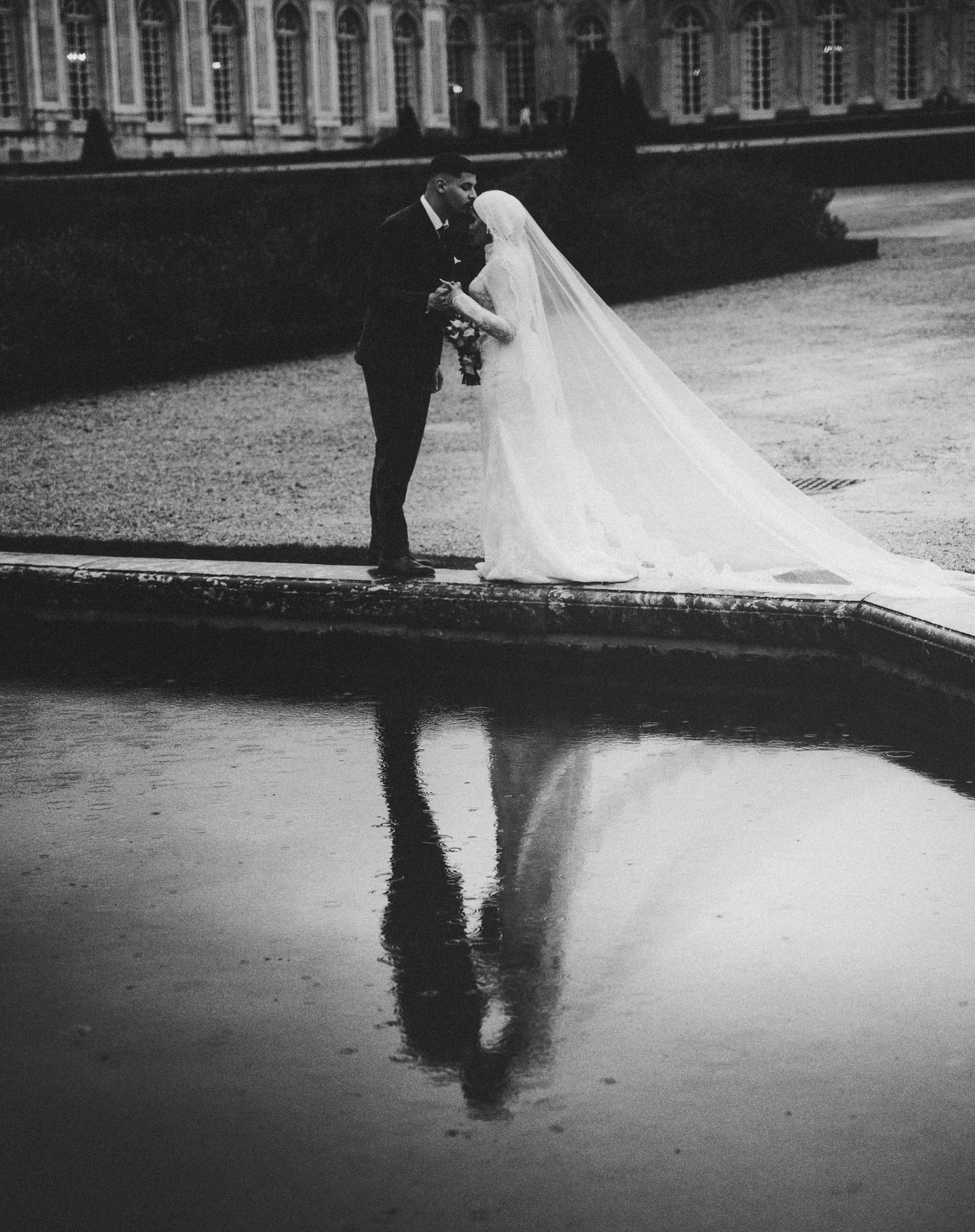 A black-and-white photo of a bride and groom holding hands and sharing a kiss outside near water, with a building in the background. Their reflection is visible in the water.
