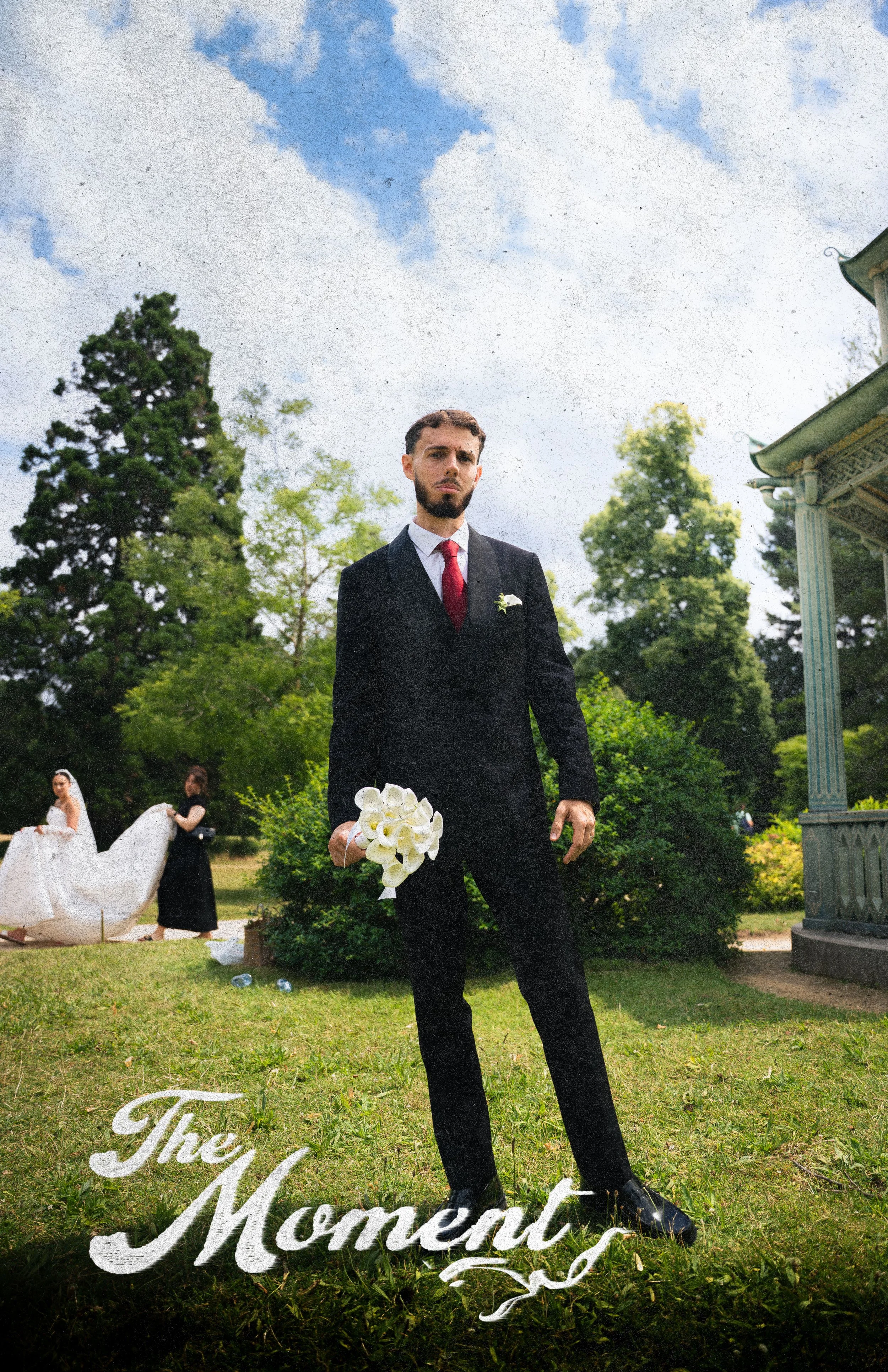A man in a suit holding a bouquet of white flowers stands outdoors on a grassy area during a wedding ceremony, with two women in wedding dresses and a decorative sculpture of a swan in the background, under a partly cloudy sky and surrounded by trees
