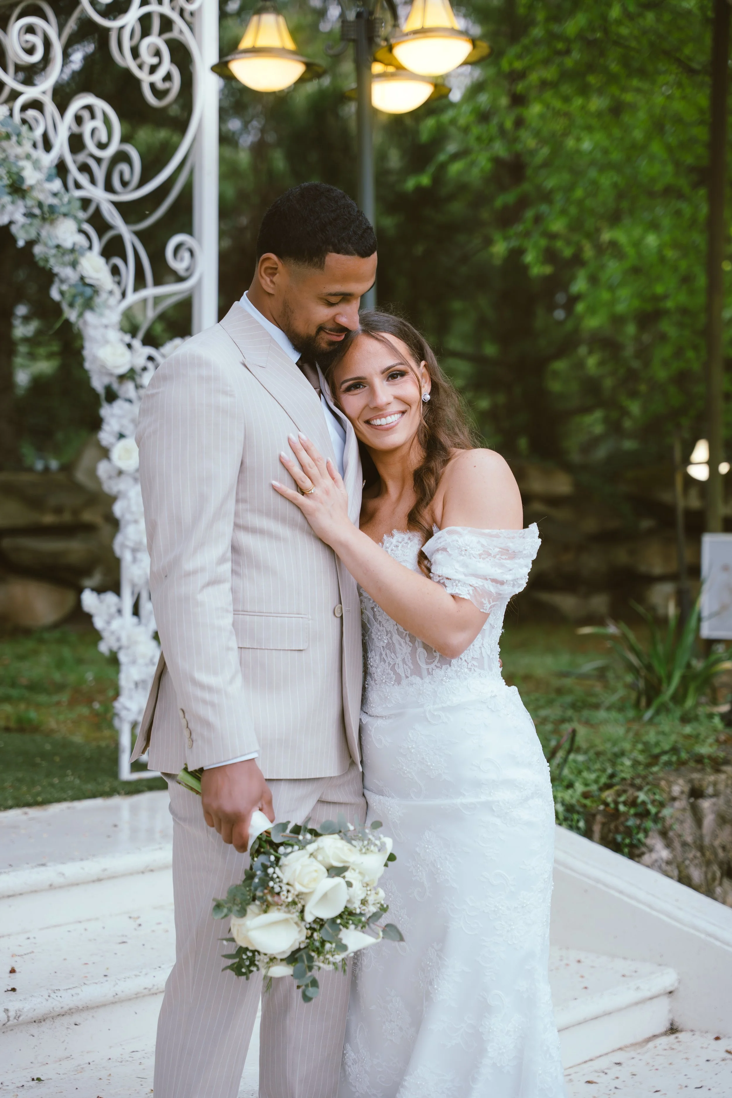 A bride and groom are embracing outdoors during their wedding, with the bride smiling at the camera and the groom looking down. The bride is in a white lace gown, and the groom in a light-colored striped suit holding a bouquet of white roses. There i