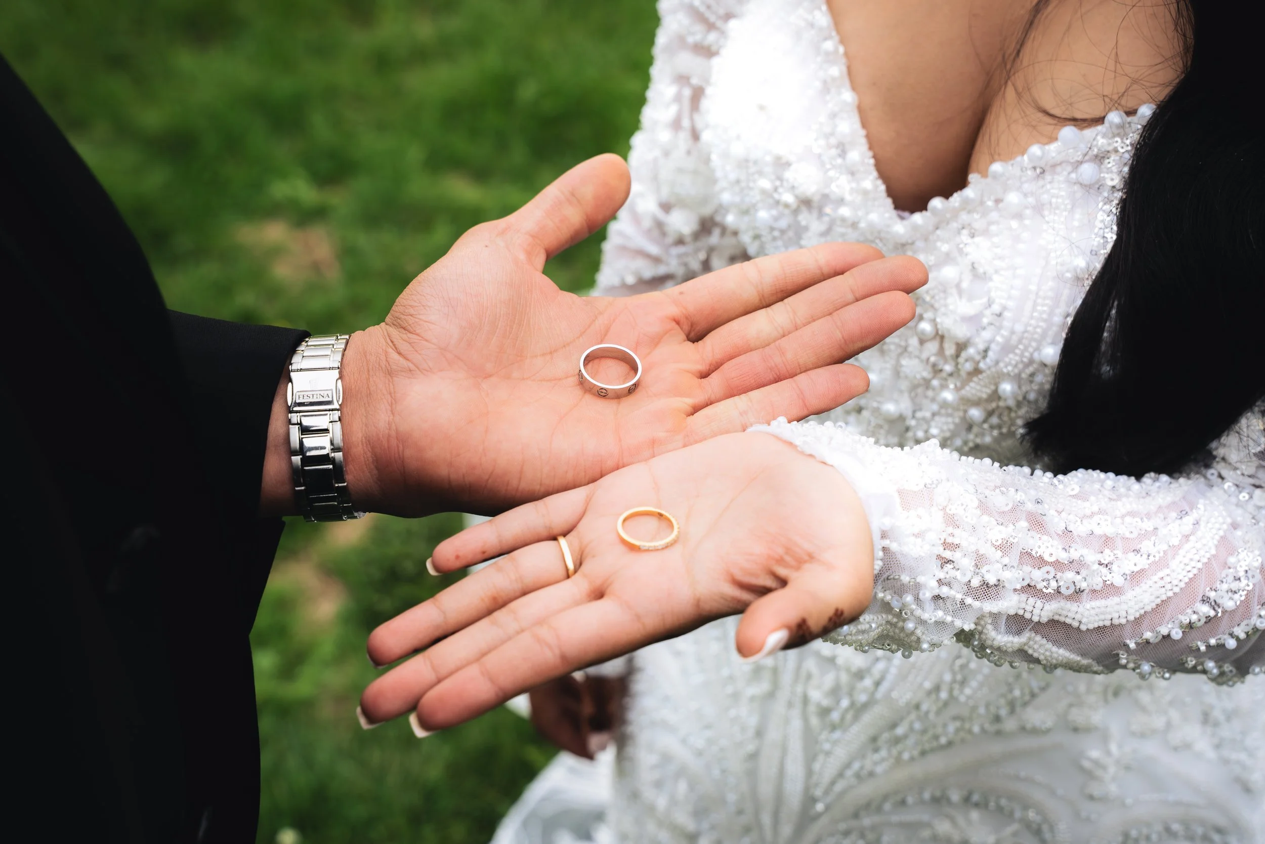 Close-up of a man and woman during a wedding ceremony, holding a wedding ring with hands. The bride has on a white dress with intricate beading and lace, and the groom is wearing a black suit with a silver watch. The background is grassy outdoors.