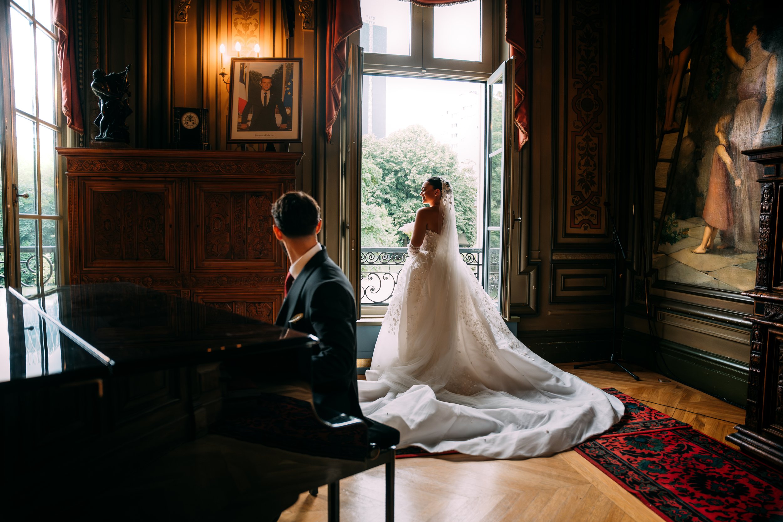 A bride in a white wedding gown standing on a balcony near a large open window, looking outside, with a man in a tuxedo sitting at a grand piano inside a decorated room. The room has ornate woodwork, a framed portrait on the wall, and artwork on the 