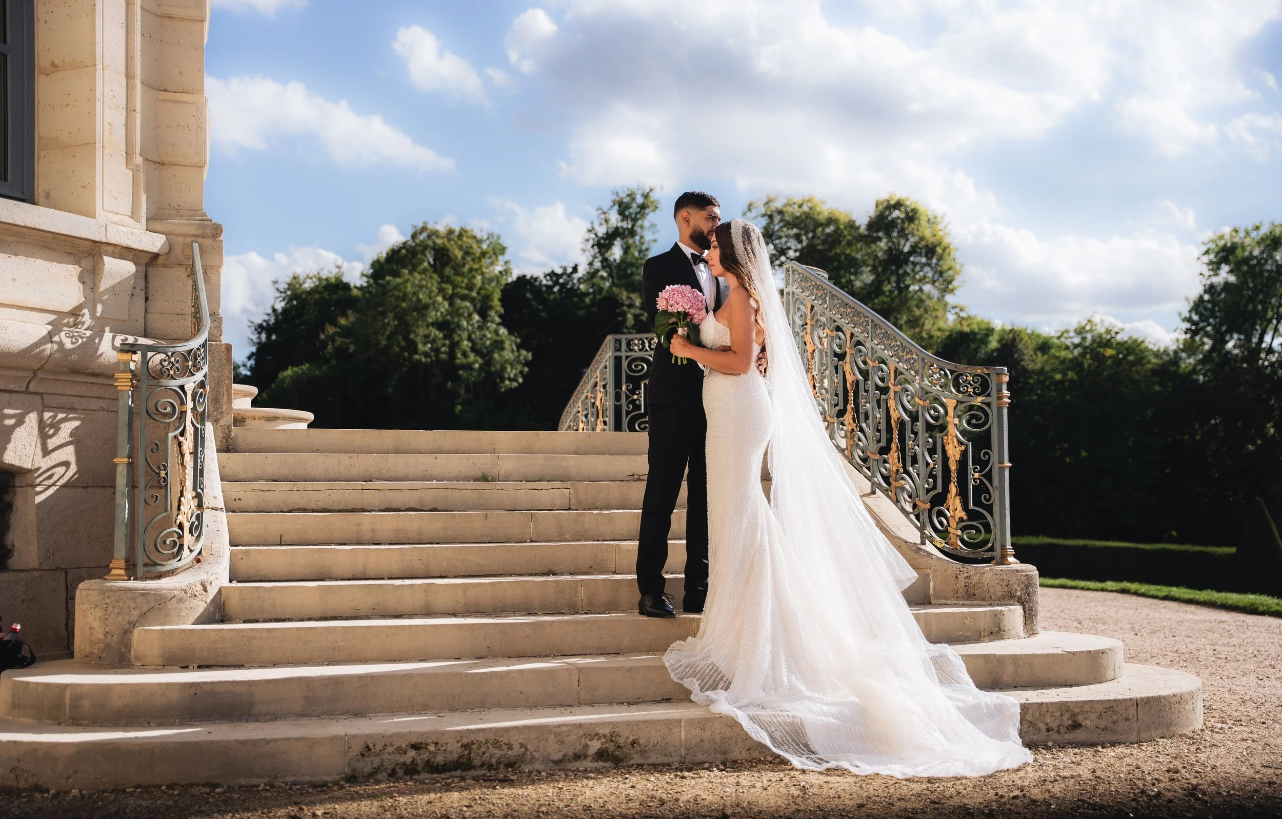 A bride and groom standing close together on outdoor stone stairs. The bride is wearing a white wedding gown and holding a bouquet of pink flowers, while the groom is in a black tuxedo. They are embracing and looking into each other's eyes, with a de