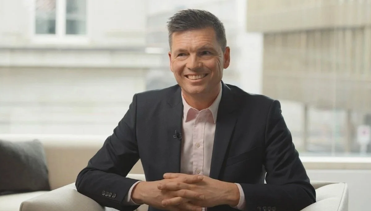 A man in a dark suit and white shirt sits on a beige couch, smiling and clasping his hands on the table in front of him, with a bright window behind him.