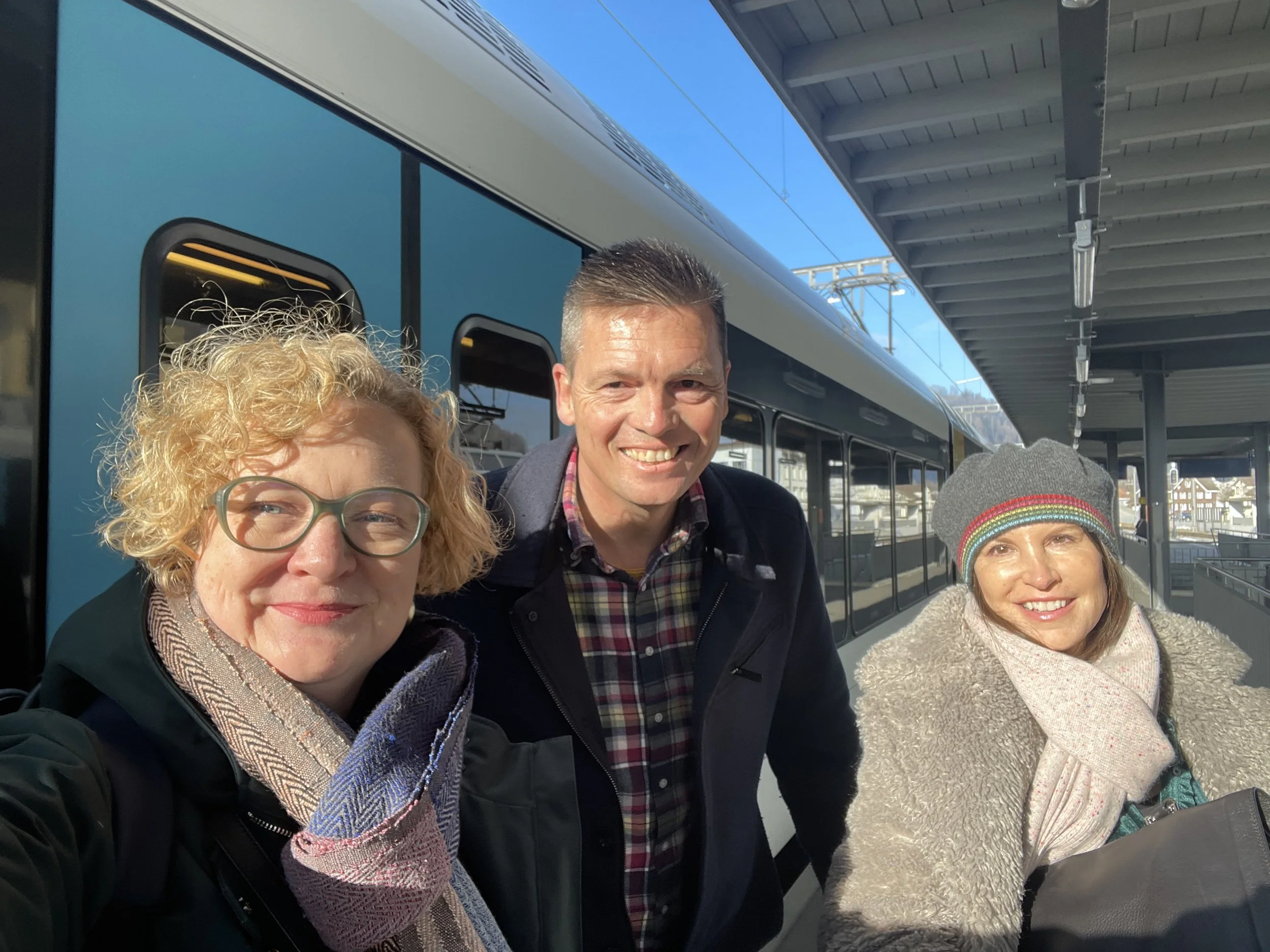 Three women and one man smiling at a train station platform under a covered awning, with a blue train in the background.