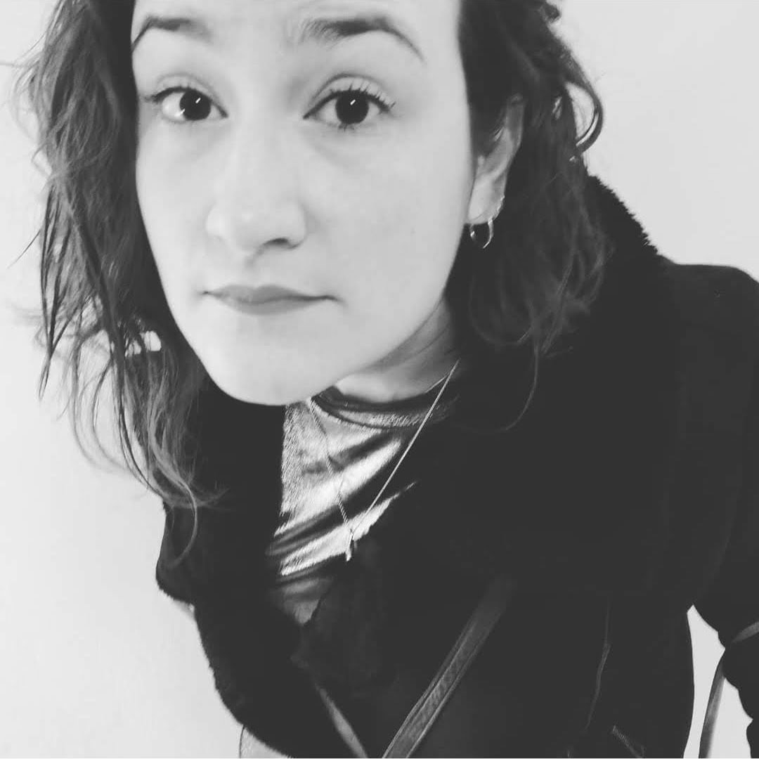 Close-up black and white photo of a woman with wavy hair, wearing earrings, a necklace, and a dark jacket, looking at the camera.