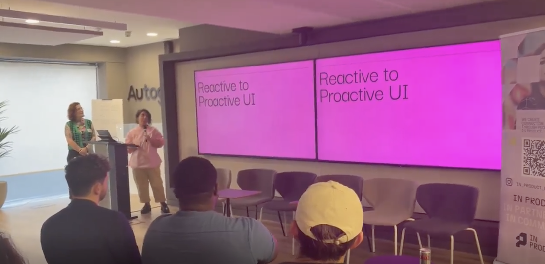 Two women are giving a presentation in a conference room with large screens displaying the text 'Reactive to Proactive UI.' Audience members are seated, watching the presentation.