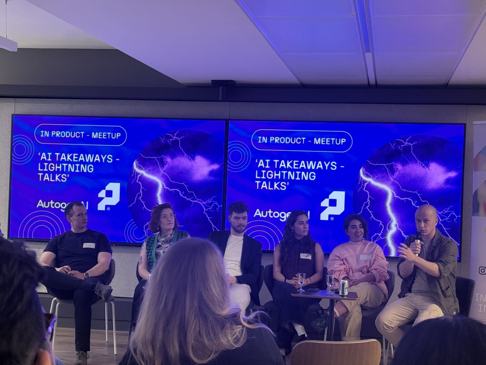 People sitting on chairs in a conference room listening to a speaker. Two screens behind them display a presentation titled 'AI Takeaways - Lightning Talks' with a lightning image. One person on the right is speaking into a microphone.