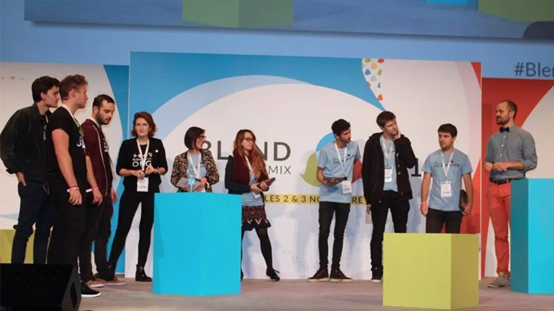 A group of people standing on a stage, some with microphones and others with tablets or papers, participating in a panel or discussion at an event with a colorful backdrop.