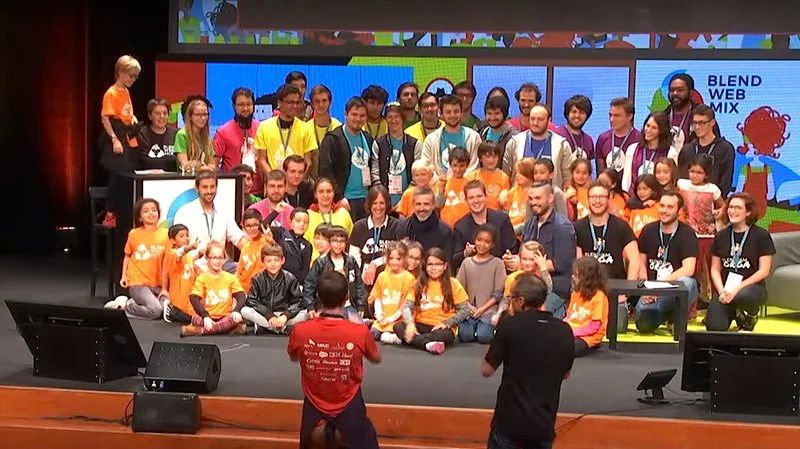 Group of children and adults on stage at a conference or event, with two photographers taking pictures in the foreground, and a colorful animated background.