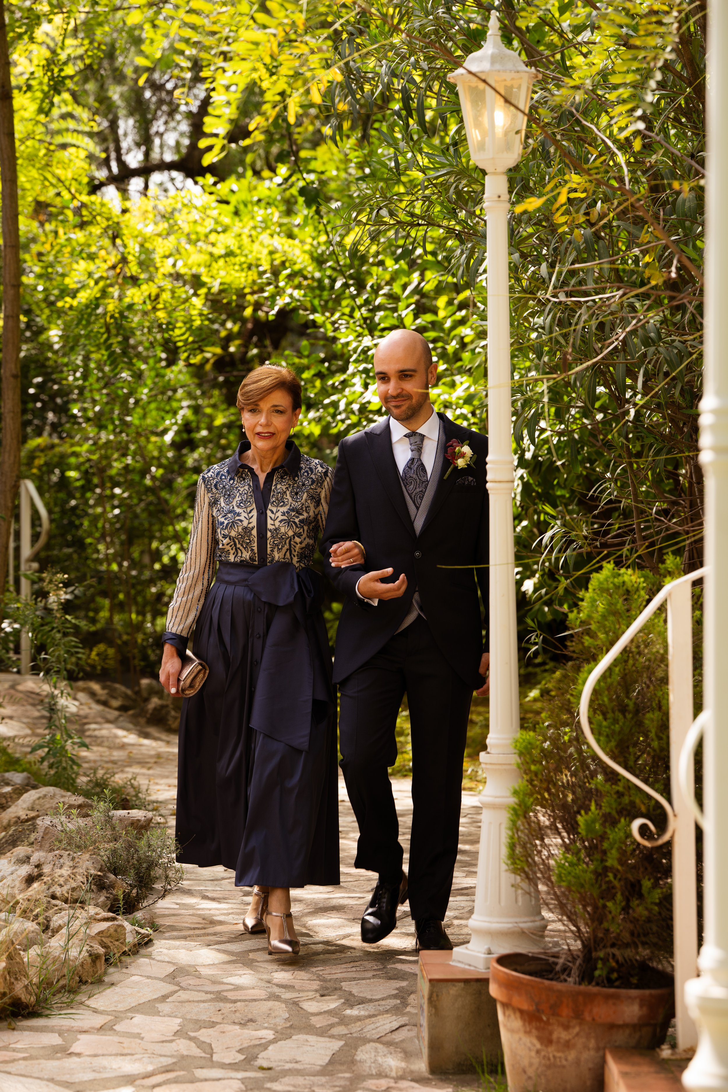 Un hombre y una mujer caminando por un sendero en un jardín, la mujer lleva un vestido largo y la mujer un traje formal, ambos sonrientes