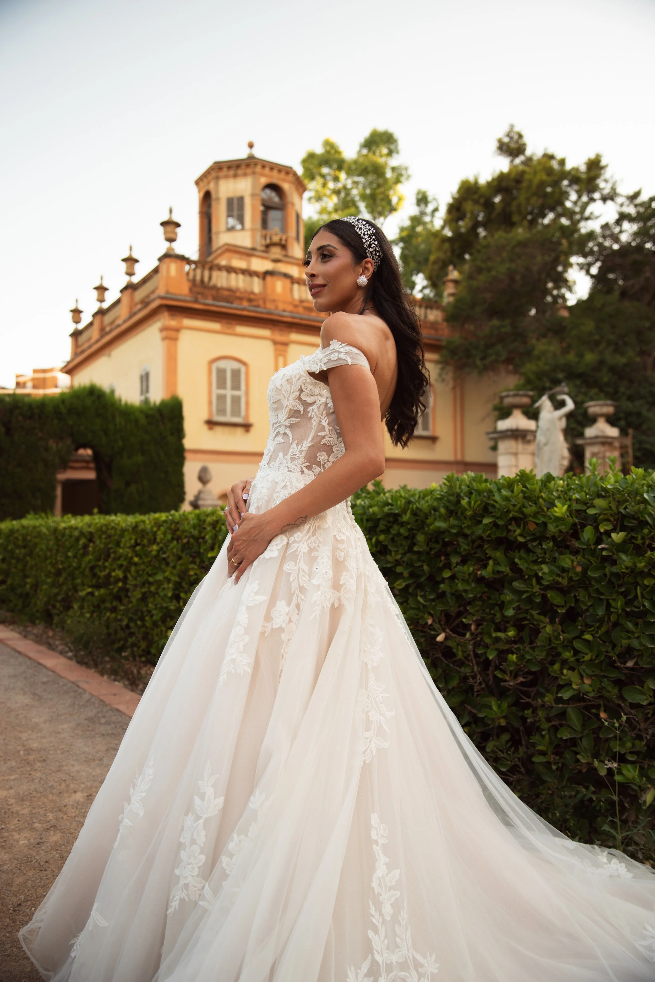 Mujer con vestido de novia de encaje y peinado con accesorios, posando al aire libre en un jardín con árboles y un edificio antiguo en el fondo.