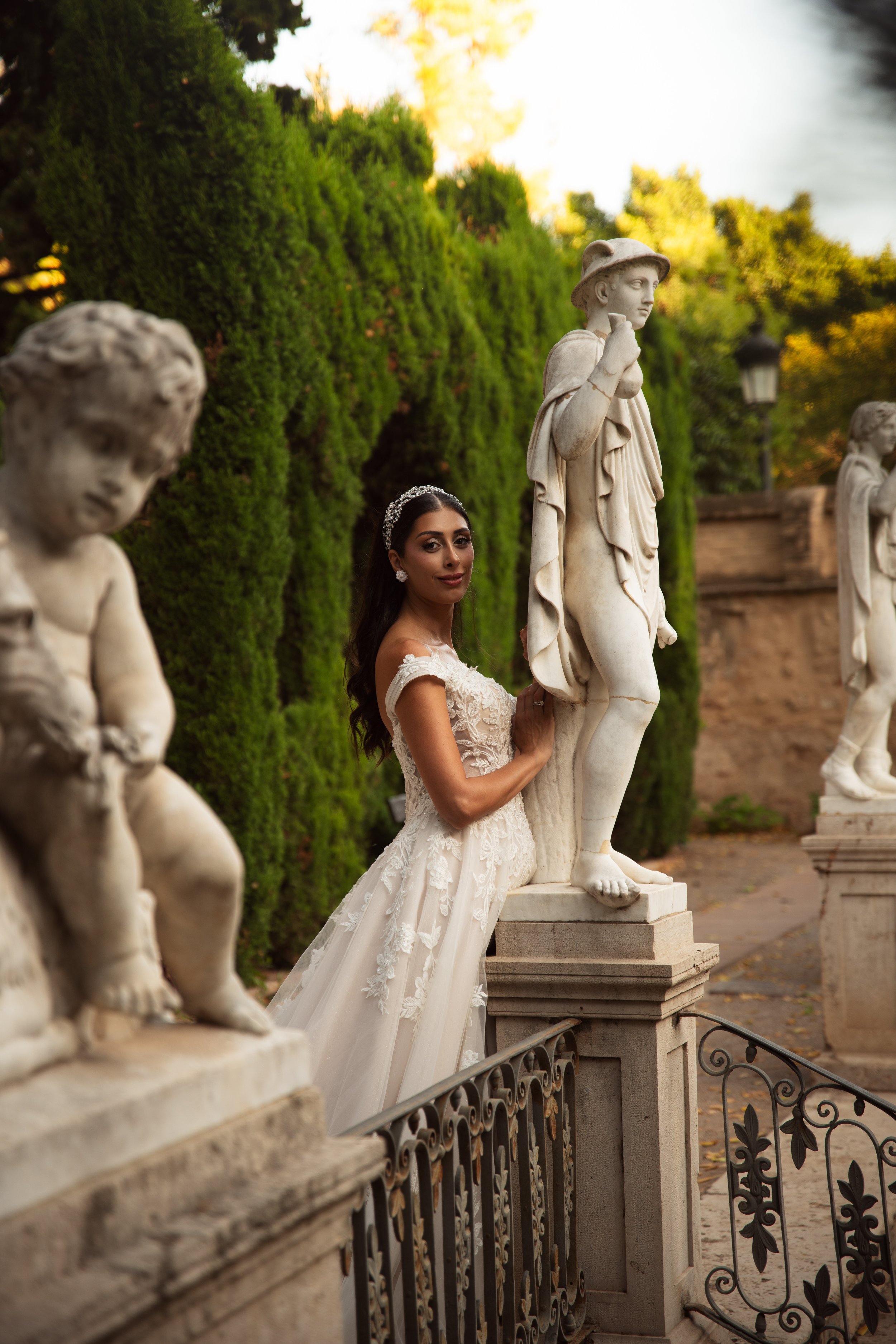 Mujer con vestido de novia sosteniendo una estatua clásica en un parque con árboles verdes y faroles en el fondo.