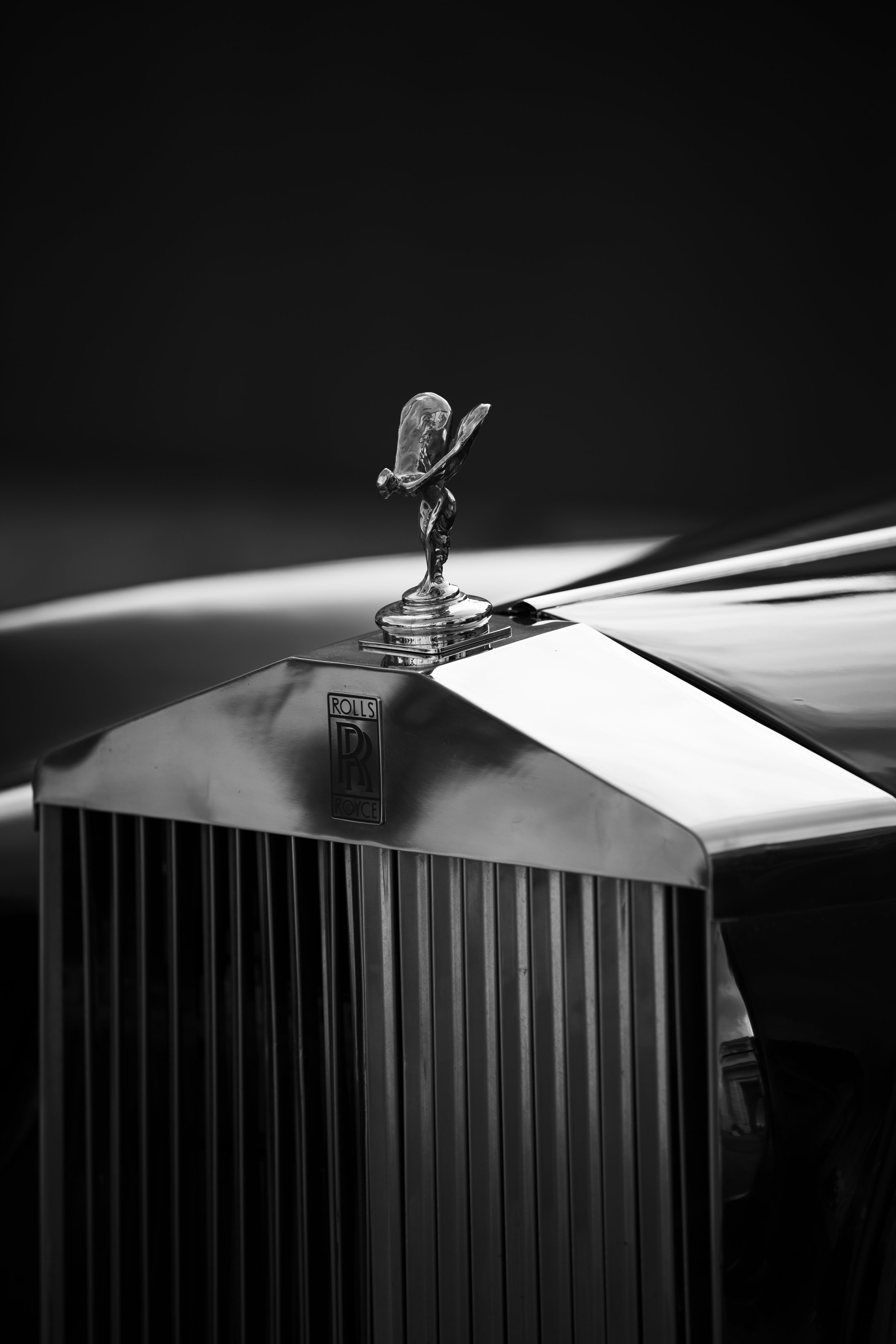 Vista en primer plano de la parrilla frontal de un automóvil clásico de lujo tipo Rolls Royce, con el emblema Spirit of Ecstasy en la parte superior, en una imagen en blanco y negro.