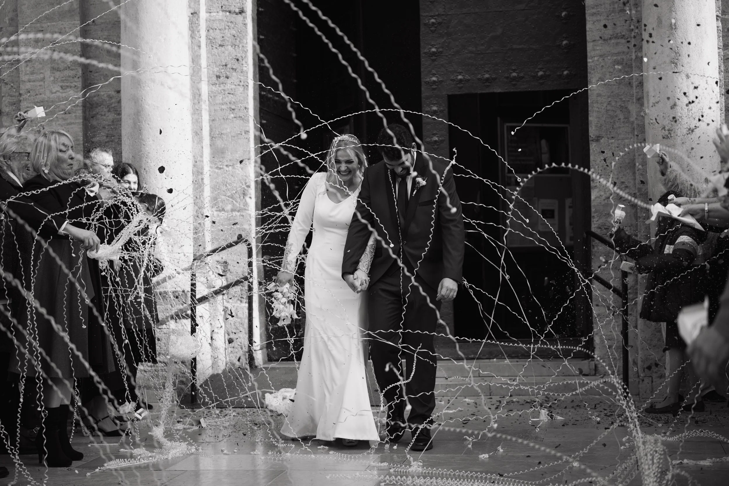 Pareja de recién casados entrando a un edificio, rodeados de invitados lanzando serpentinas durante la celebración de boda.