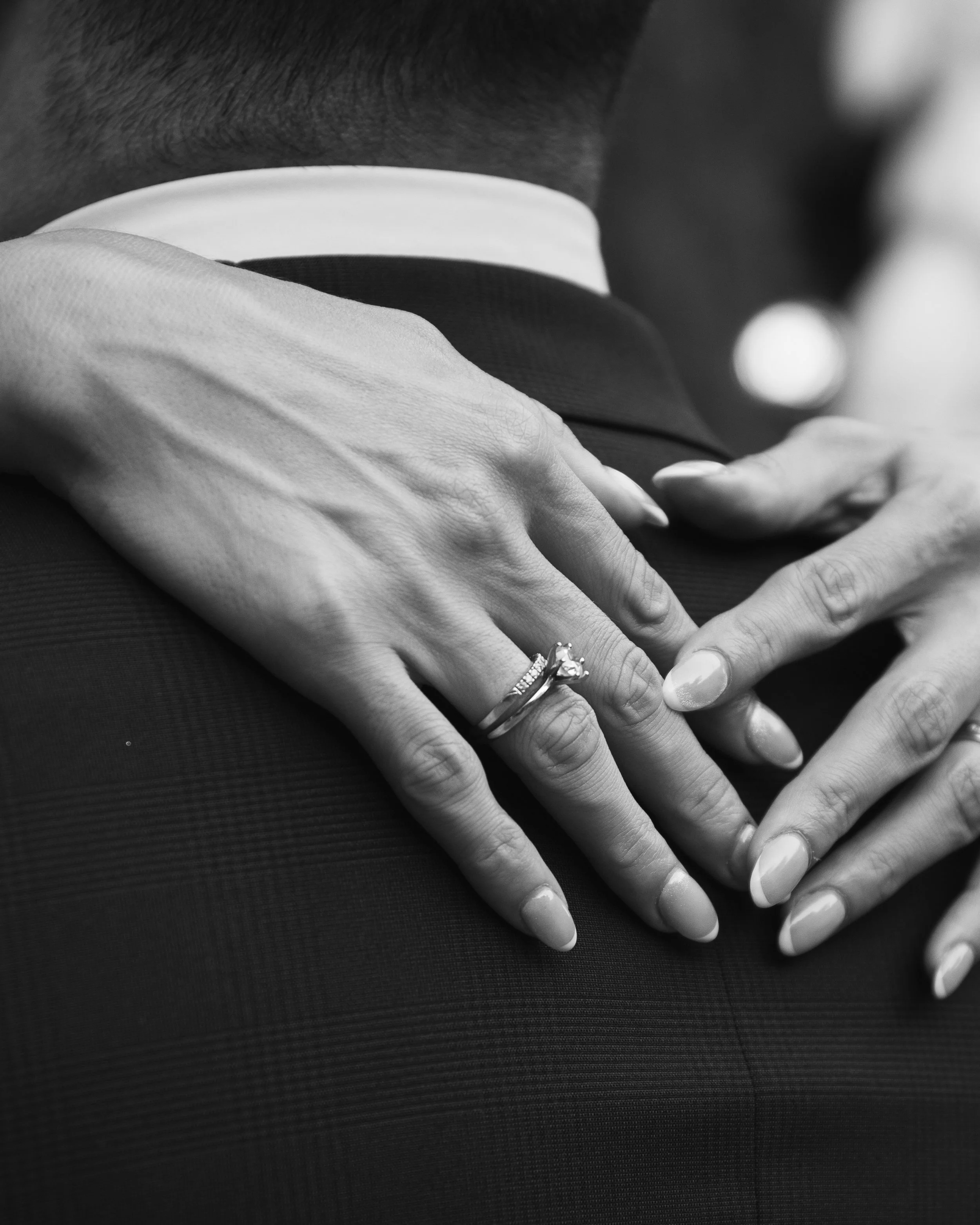 Dos manos, una con anillo de compromiso, descansando sobre la espalda de otra persona, en un retrato en blanco y negro.