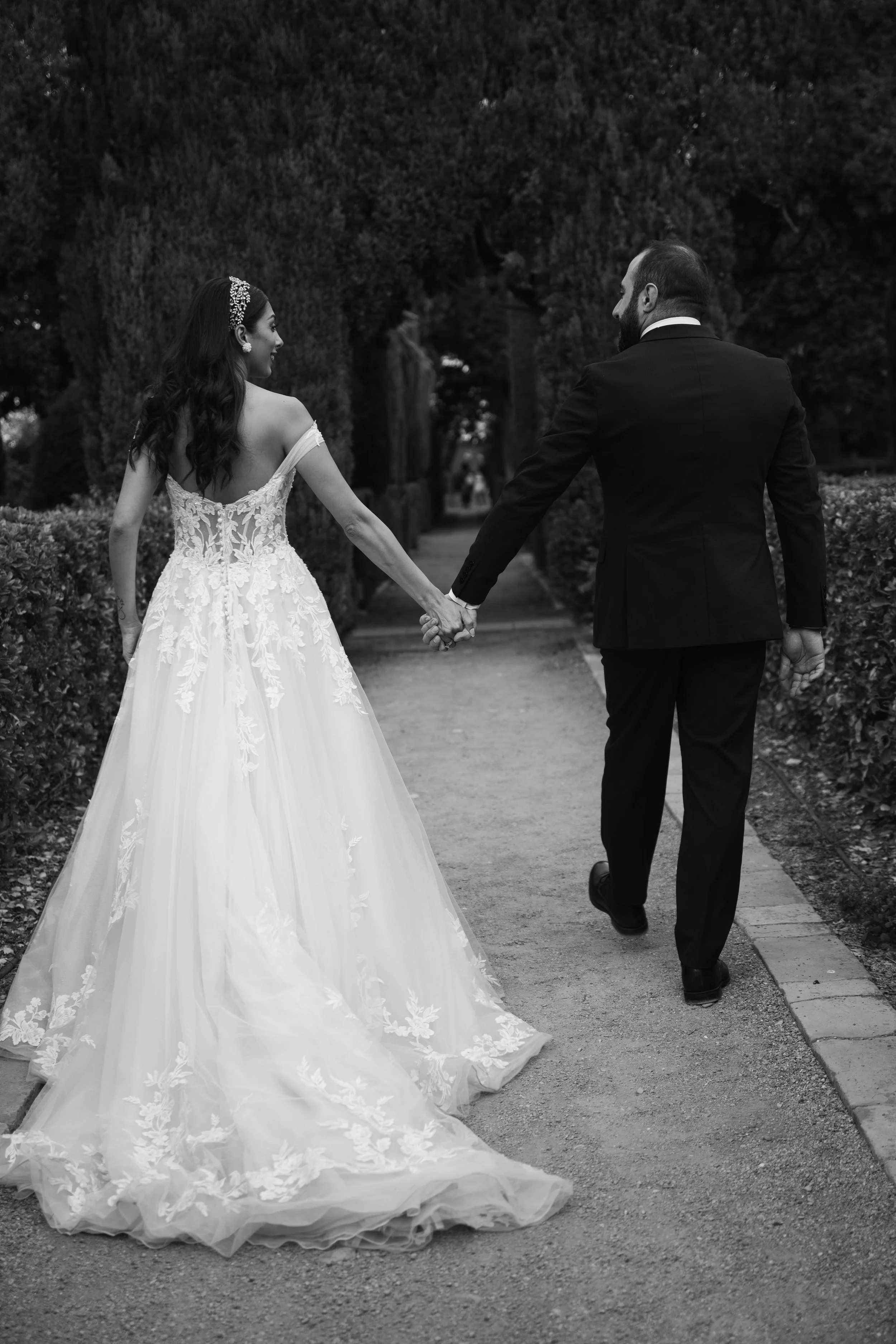 Pareja de novios caminando por un sendero con arbustos y árboles, la novia viste un vestido de novia largo y el novio lleva un traje negro, se toman de la mano y se ven felices.