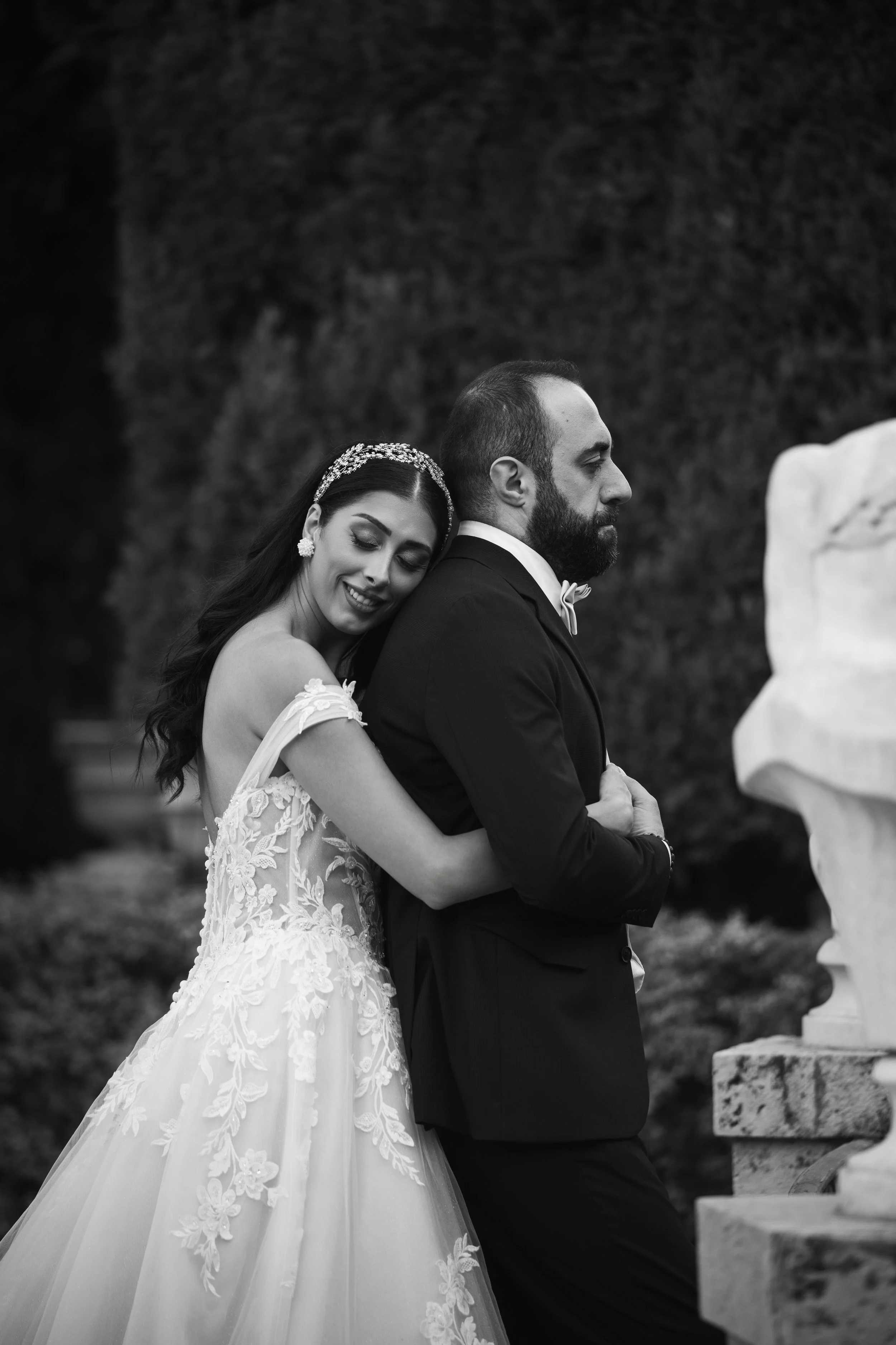 Pareja de novios en boda, la novia abraza al novio en la espalda, vestido de novia con encaje y tiara, el novio con traje de etiqueta, fondo de árboles y escultura de piedra.