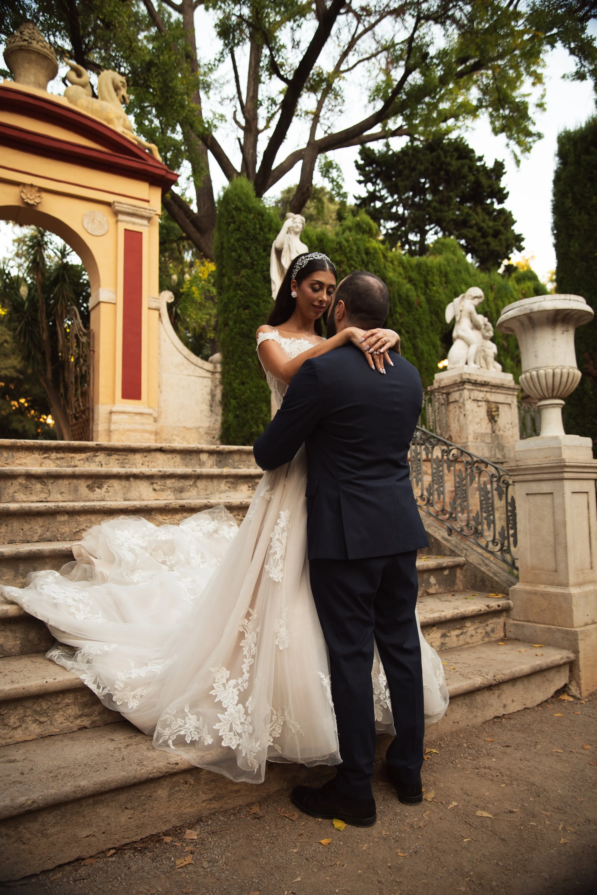 Una pareja vista de frente en una boda, la mujer lleva vestido de novia y el hombre traje negro. Están en las escaleras de piedra de un lugar decorado con estatuas y vegetación, en un ambiente al aire libre.