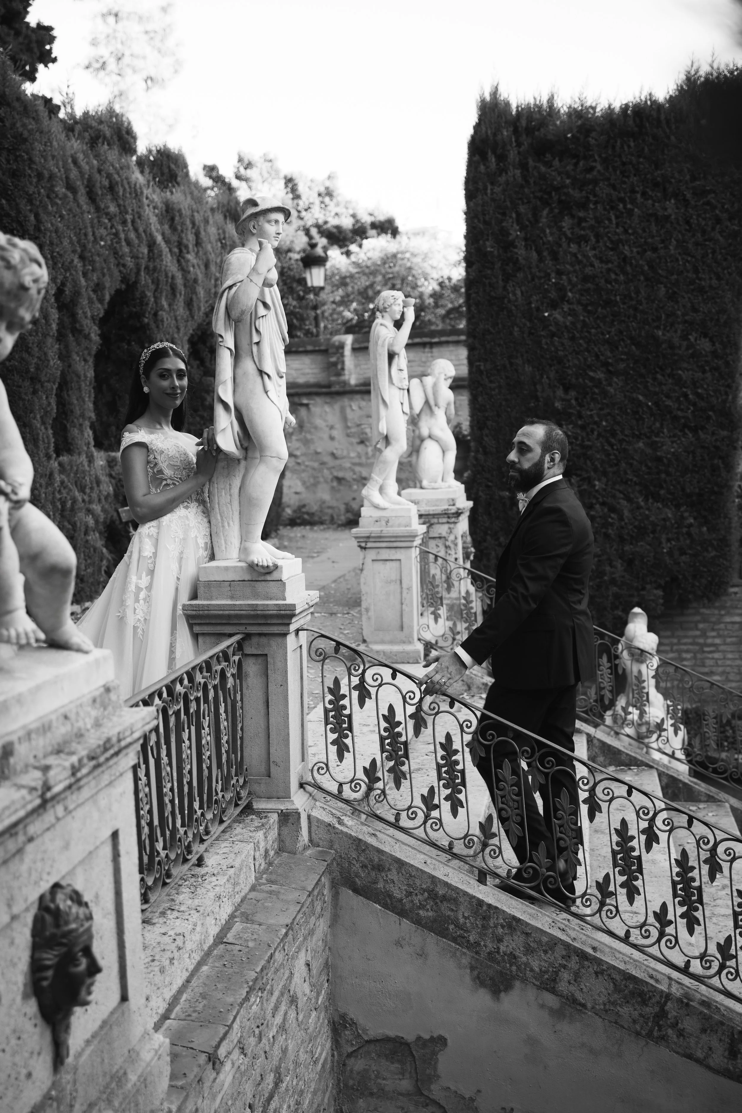 Hombre con traje de etiqueta en un jardín con estatuas de mármol y una cerca de hierro decorativo, en un entorno con árboles grandes y una fuente al fondo.