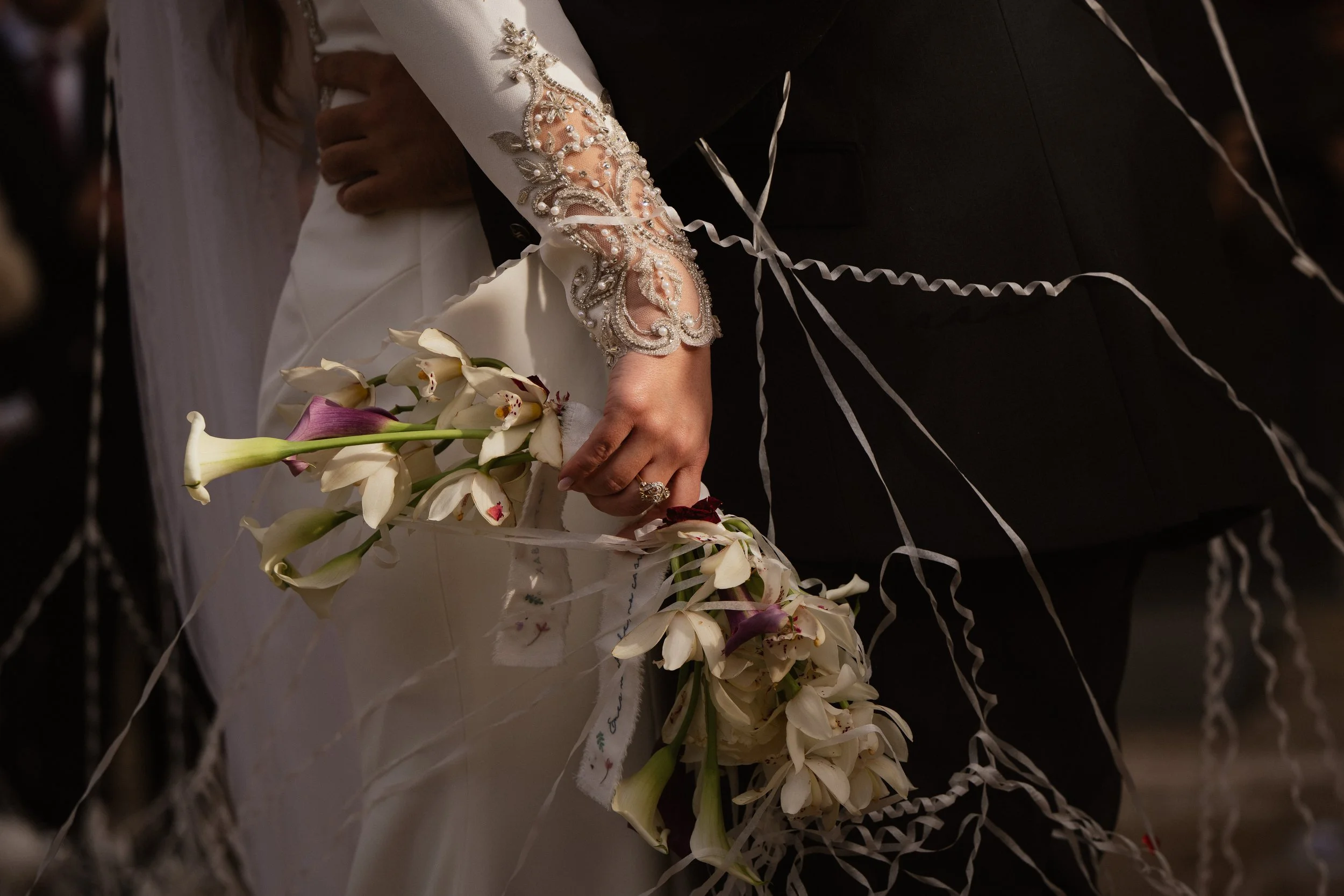 Pareja envestida en una boda, sosteniendo un ramo de flores blancas y rosas, con detalles en su vestimenta. La mujer lleva una prenda con bordados y la mano con un anillo.